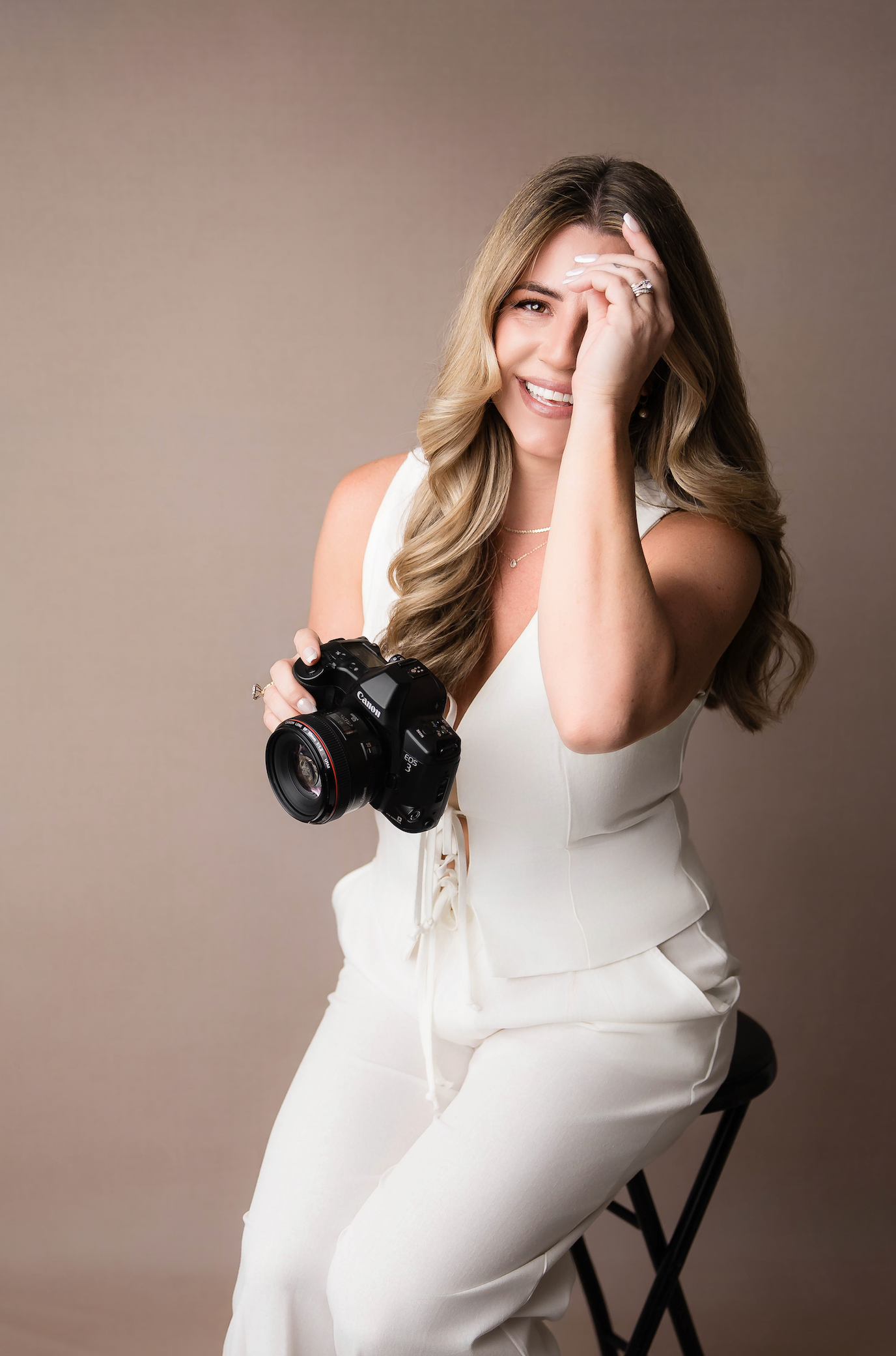 A photographer in a white outfit sits on a stool holding a DSLR camera while laughing and covering face with hand.