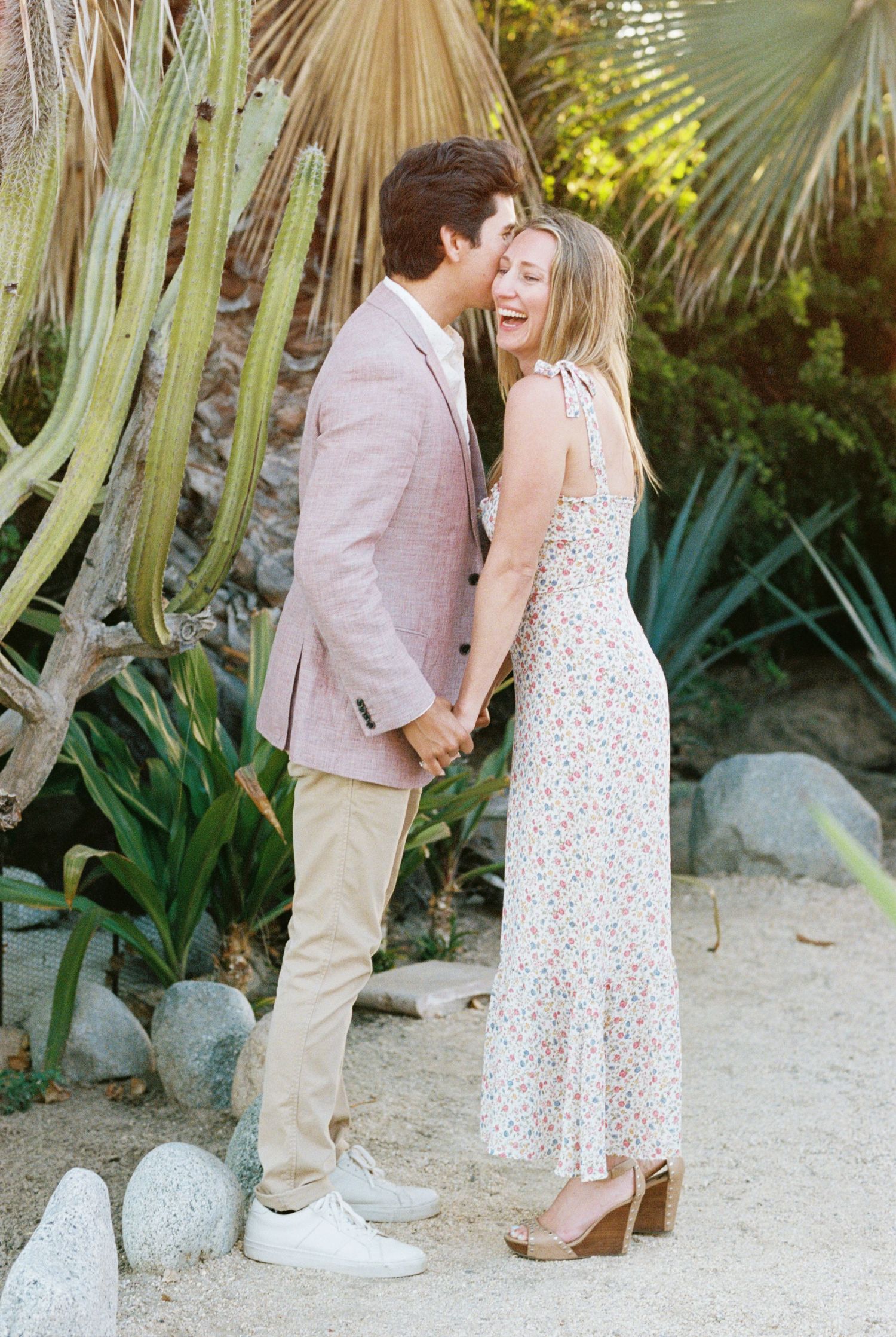 Romantic couple embraces in a garden with palm trees, wearing light summer attire for an engagement photo shoot. Kirstie Whitaker Photograph