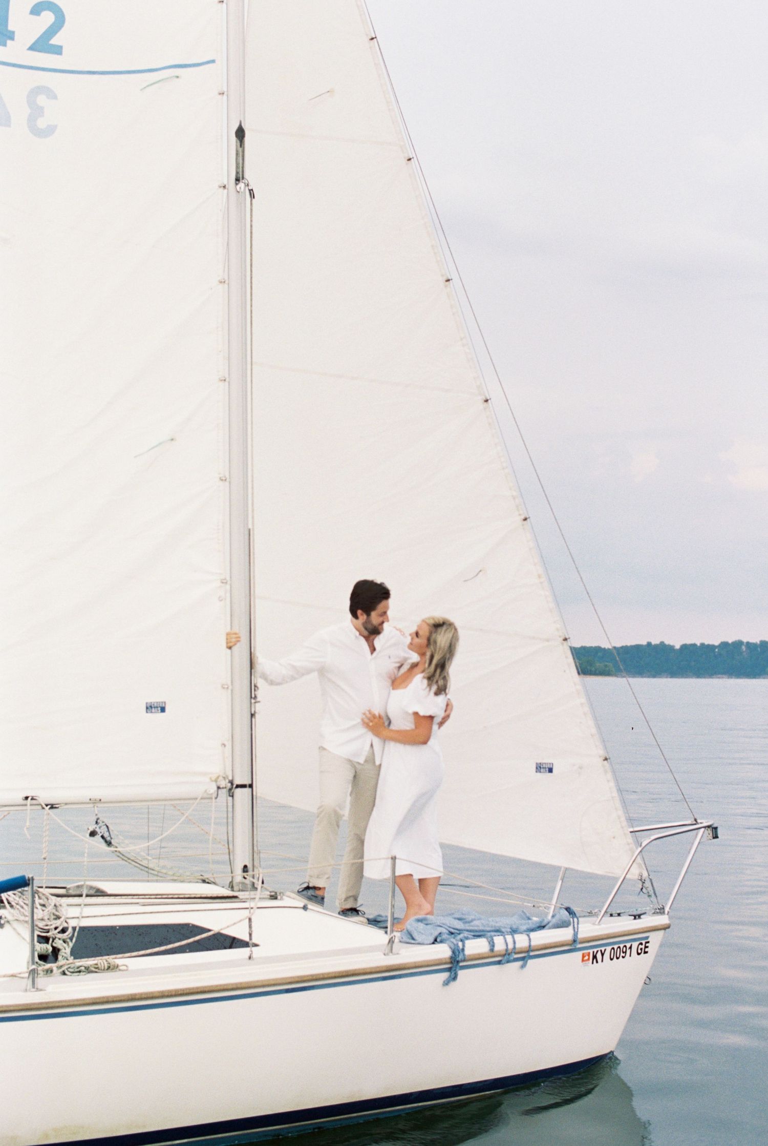 A couple in white outfits stand together on a sailboat against a calm lake backdrop on a cloudy day.