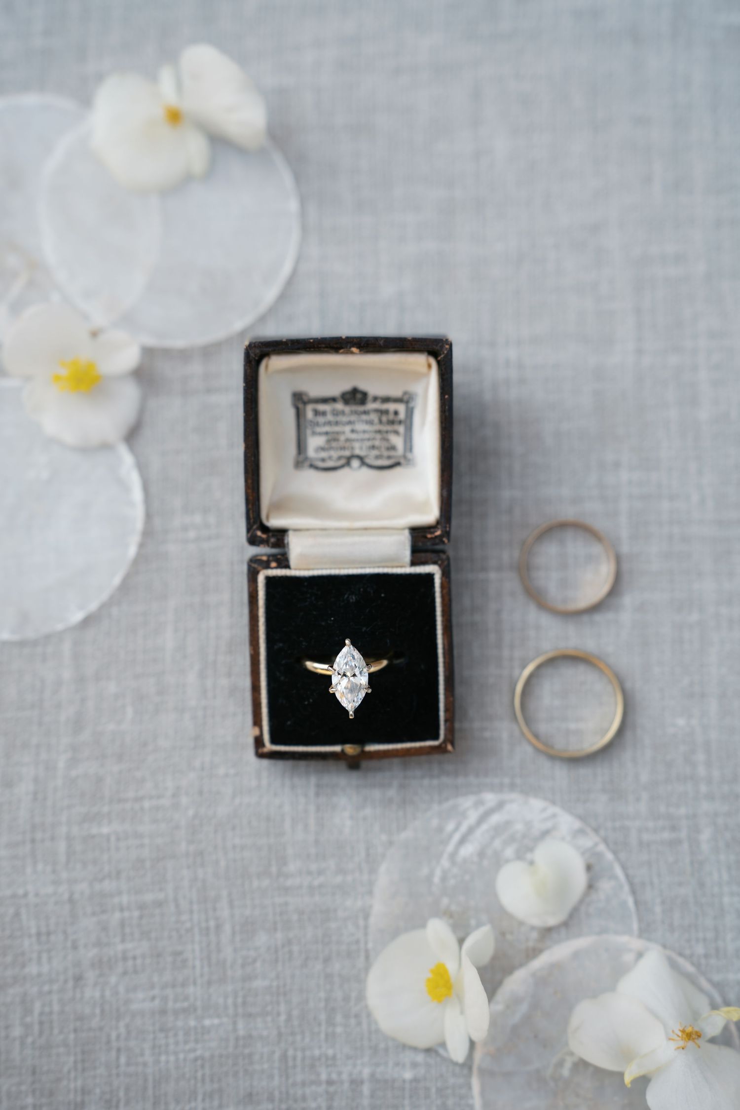 Vintage ring box with diamond engagement ring displayed on gray linen fabric with scattered white orchid petals.