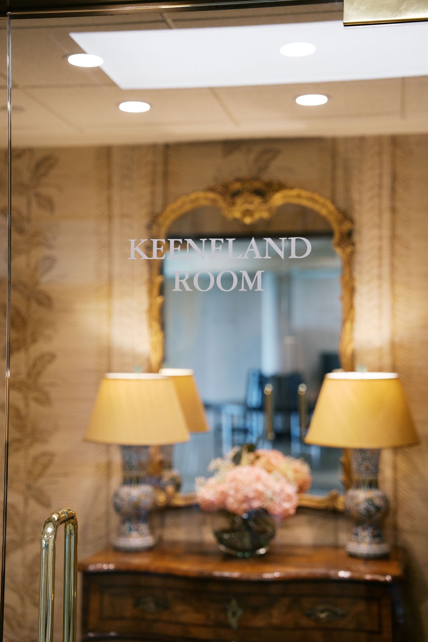 Elegant Keeneland Room entrance with ornate gold mirror, decorative lamps, and pink floral arrangement on wooden console table.