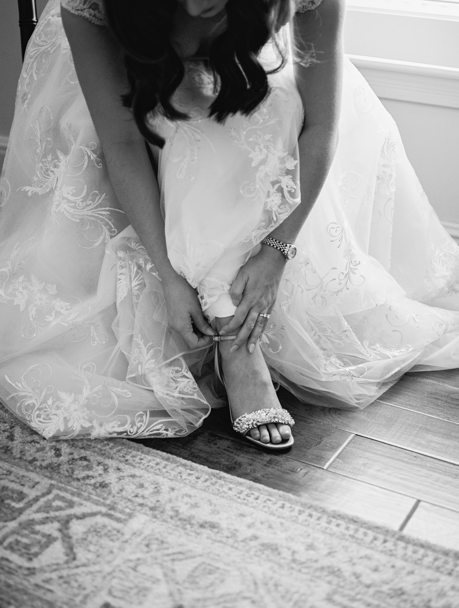 Black and white photo of a bride adjusting her sparkly sandal while wearing a flowing lace wedding dress.