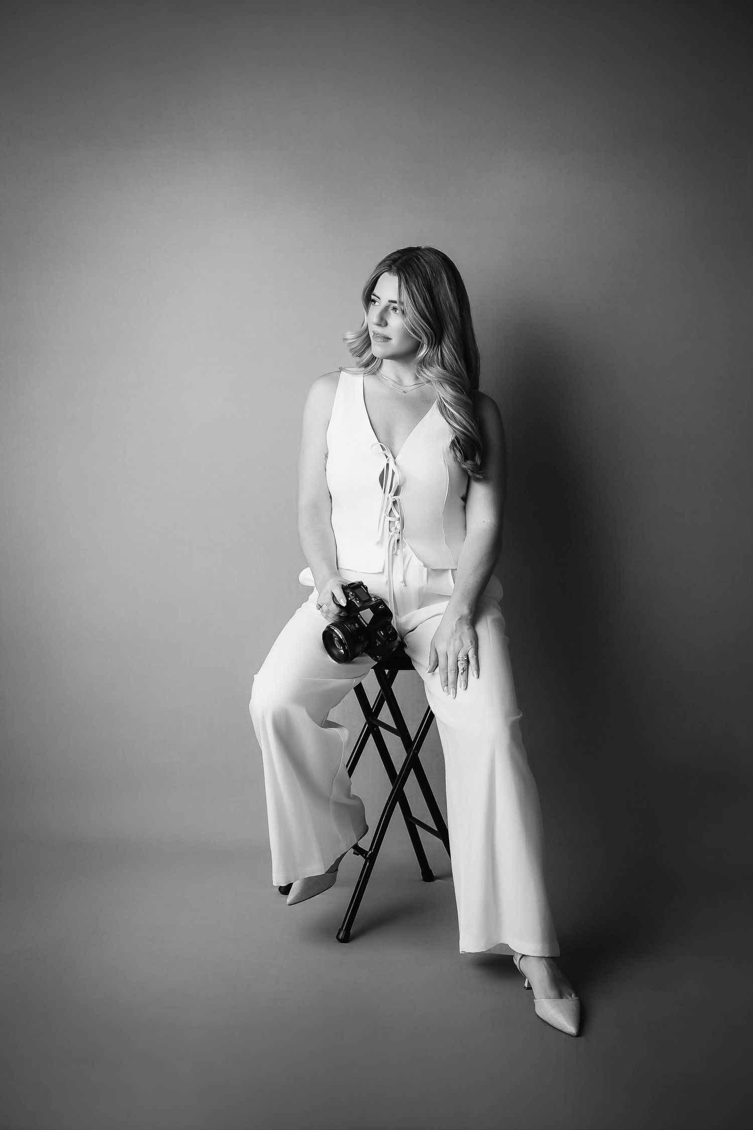 Black and white portrait of someone seated elegantly on a chair in a flowing white dress, holding a camera.