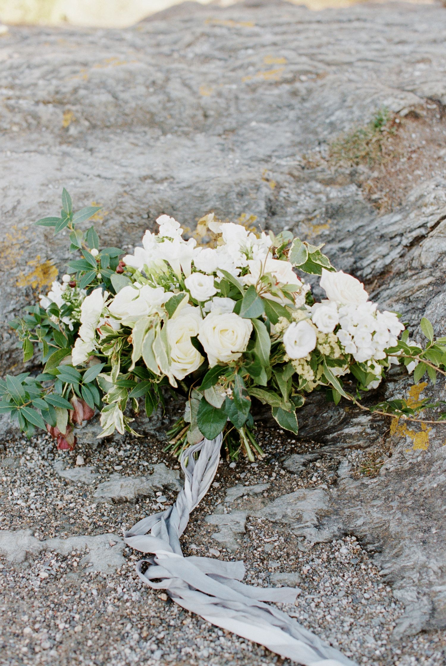 Elegant white rose and eucalyptus wedding bouquet resting on textured gray rocks with trailing silk ribbon.