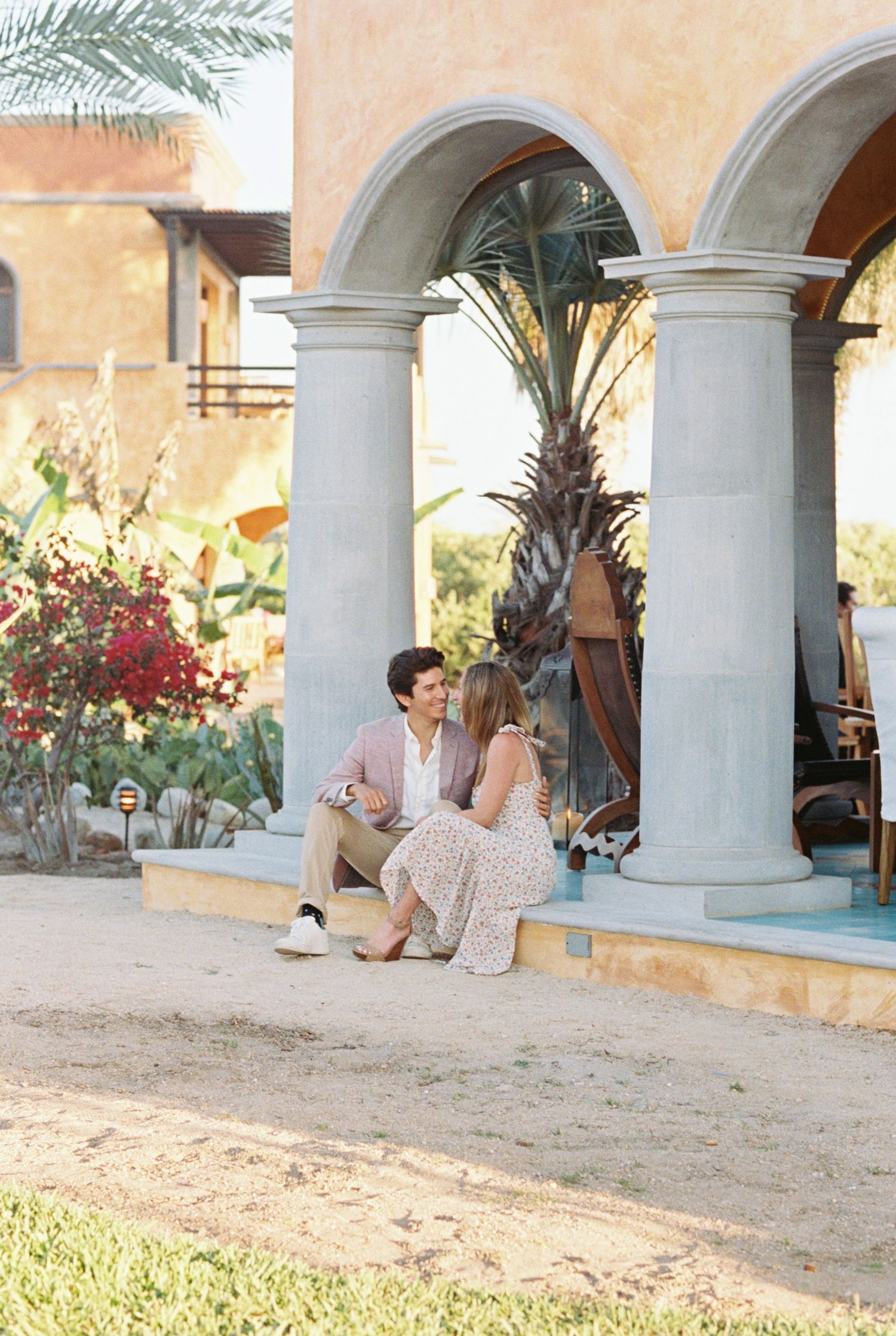 A couple sits together on the steps of a Mediterranean-style building with white columns and palm trees in the background.