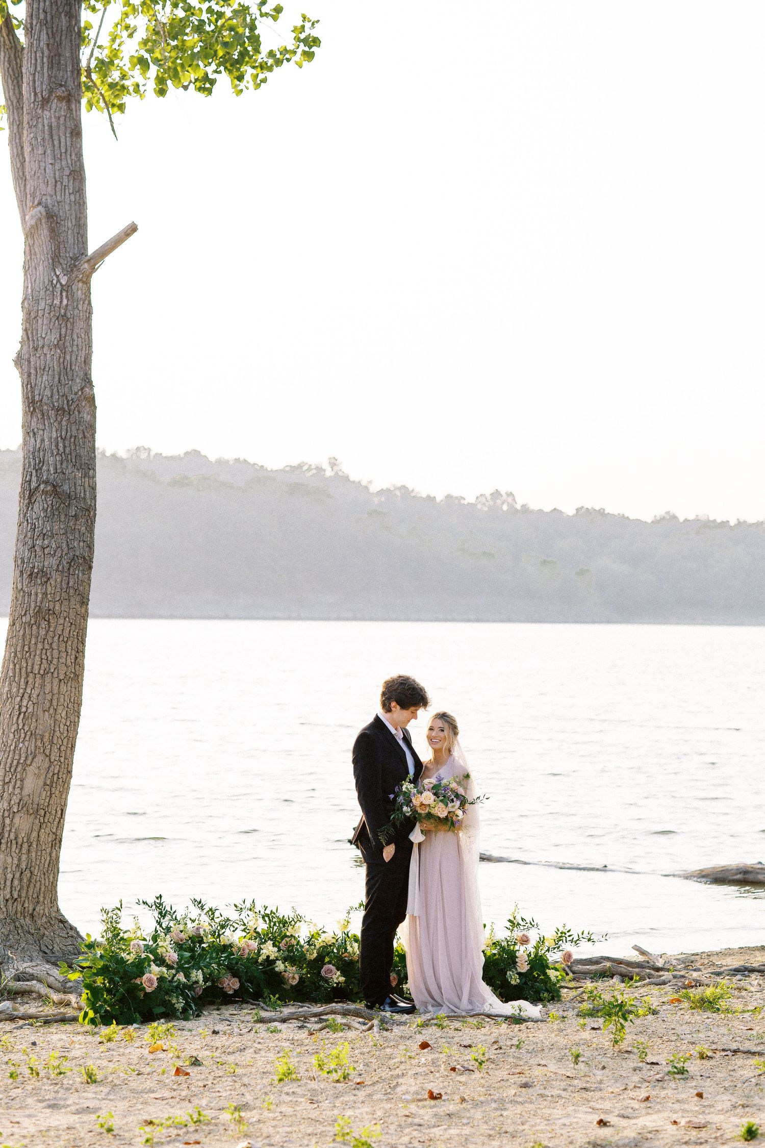 A romantic lakeside wedding scene with floral arrangements by a tree, overlooking calm waters at sunset.