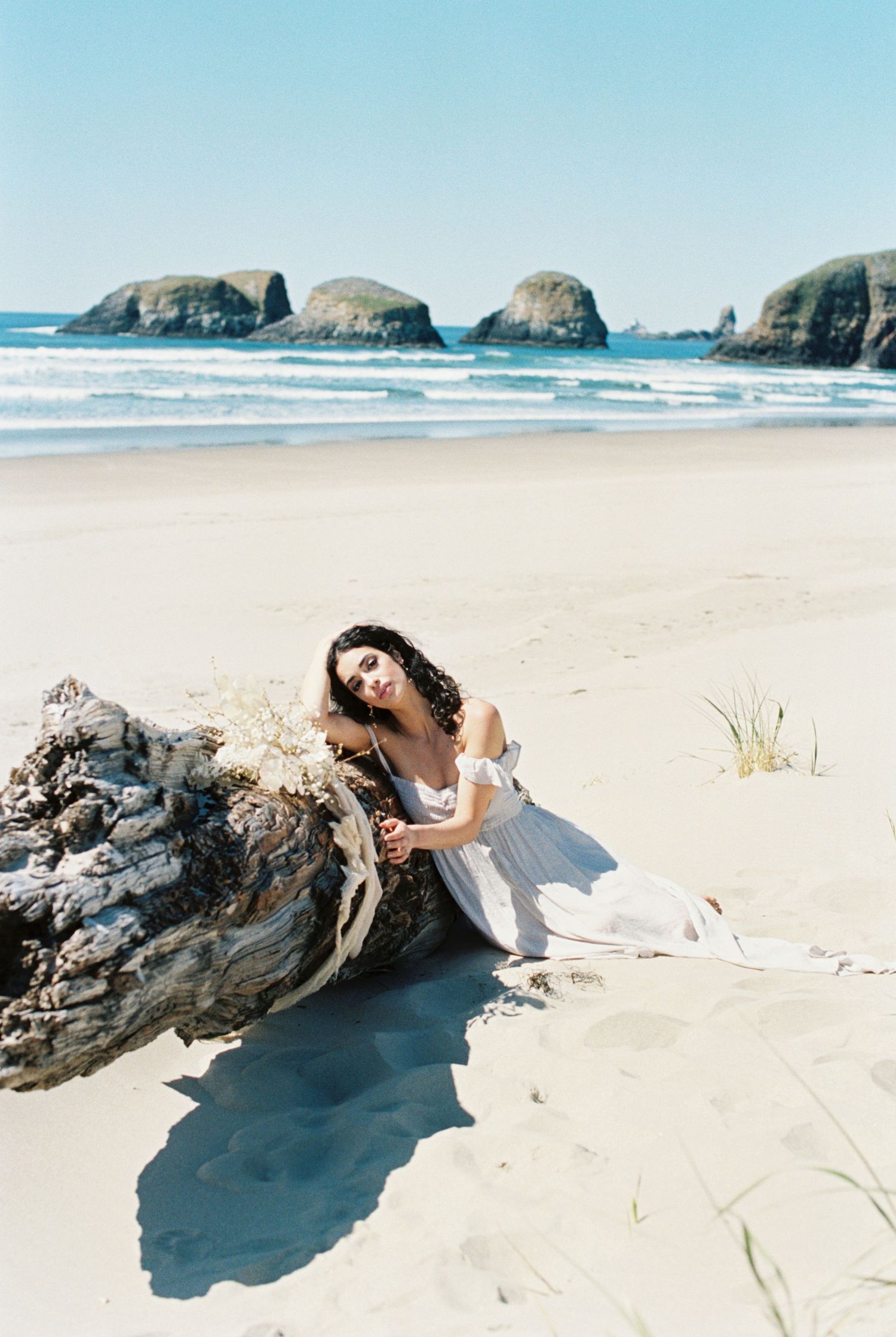 White sand beach with large rocky formations in the ocean and someone in a flowing white dress resting against driftwood.