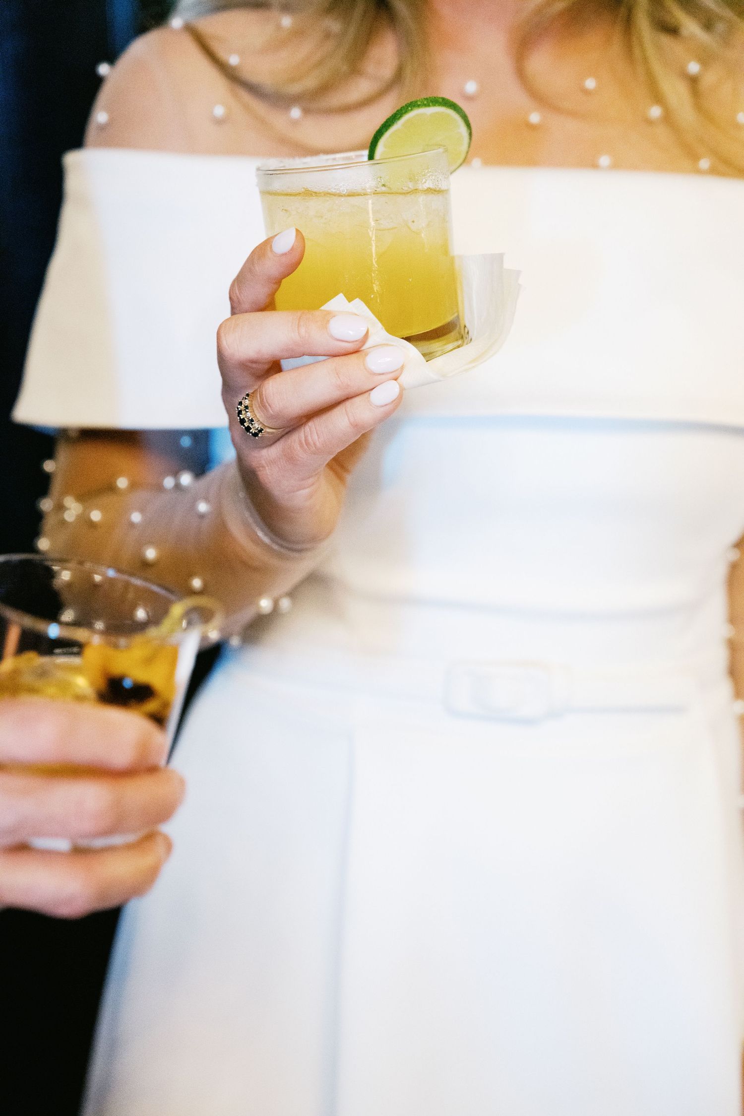 Three hands holding yellow cocktails with lime garnishes against a soft blurred background at a party or celebration.