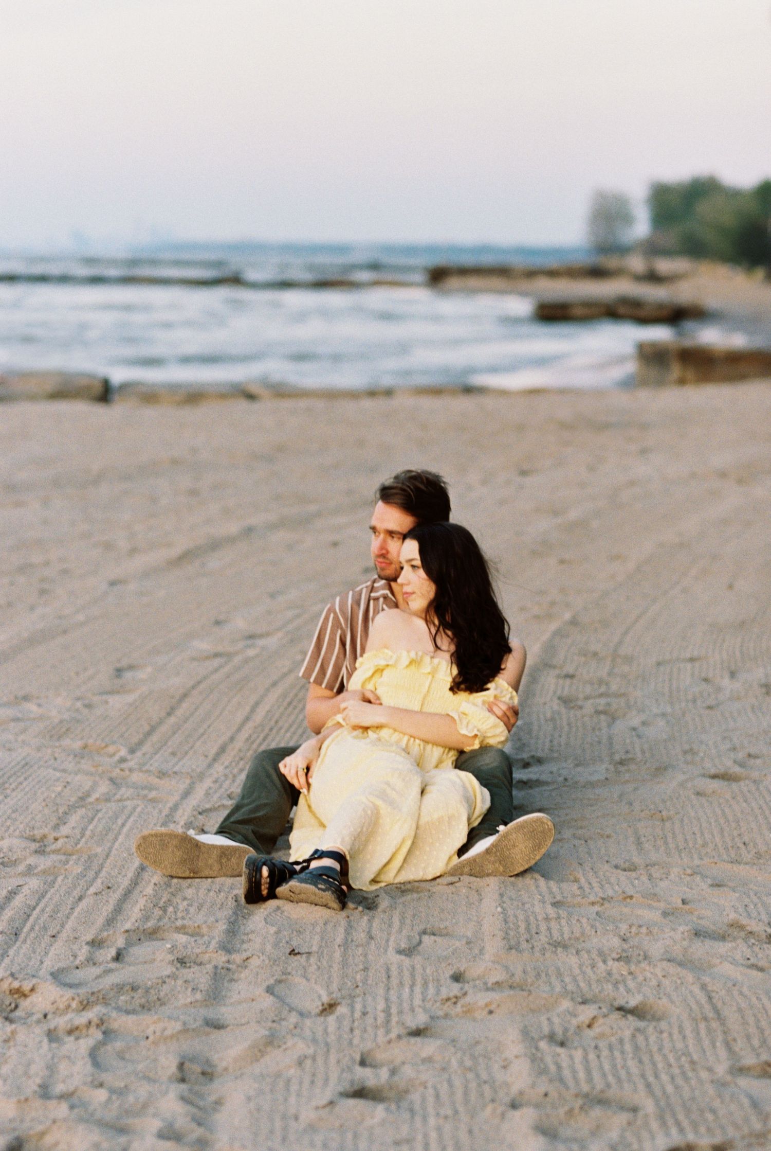 Two people share an intimate moment sitting together on a sandy beach at sunset with ocean waves and rocks in the background.