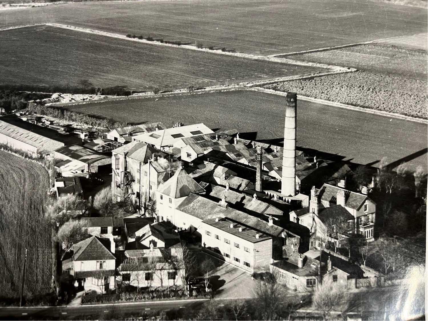 Aerial black and white photo of a historic industrial town with a tall chimney stack, factory buildings, and surrounding farmland.