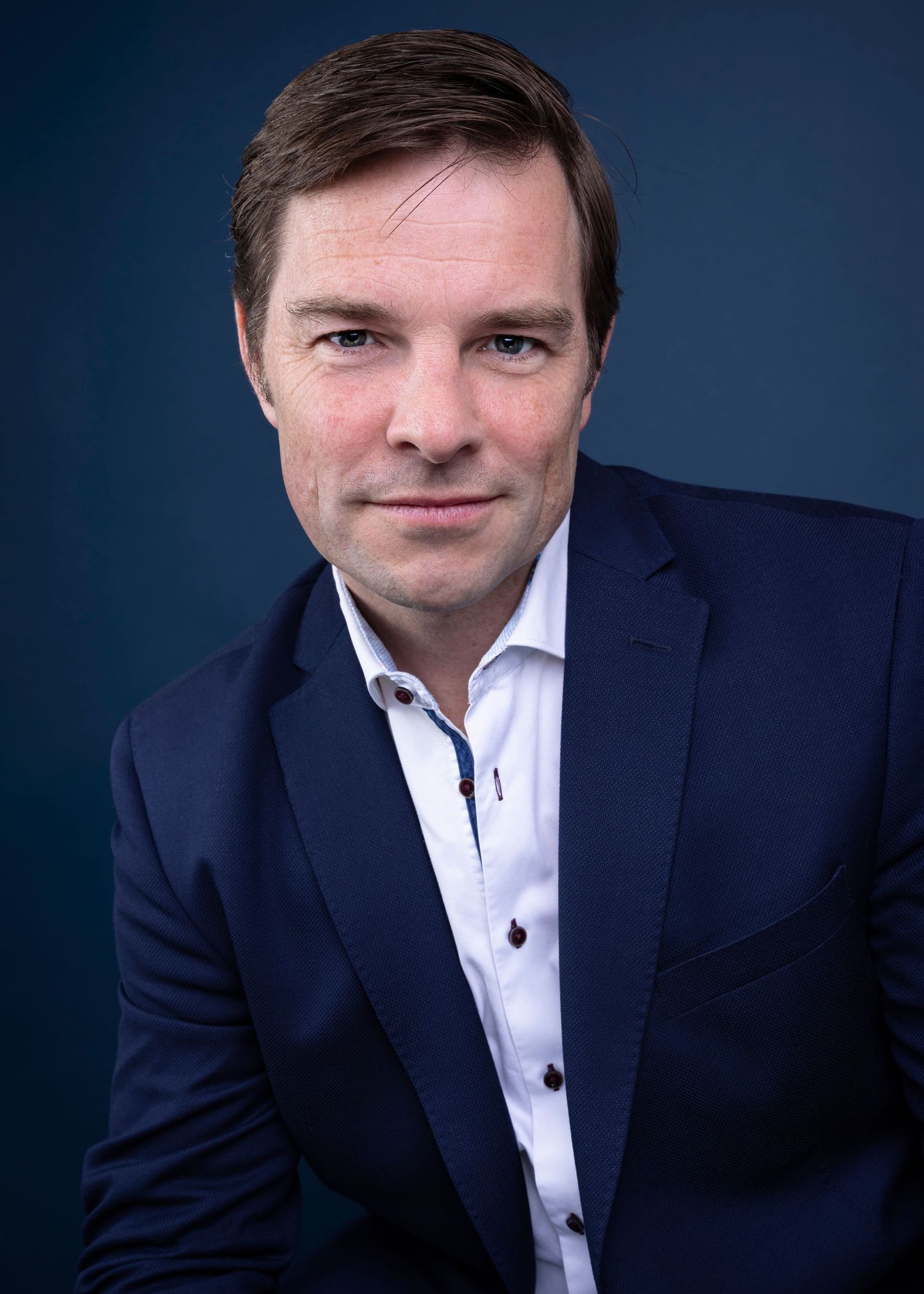 Professional headshot of a man in a navy blue blazer and white shirt against a dark blue background.
