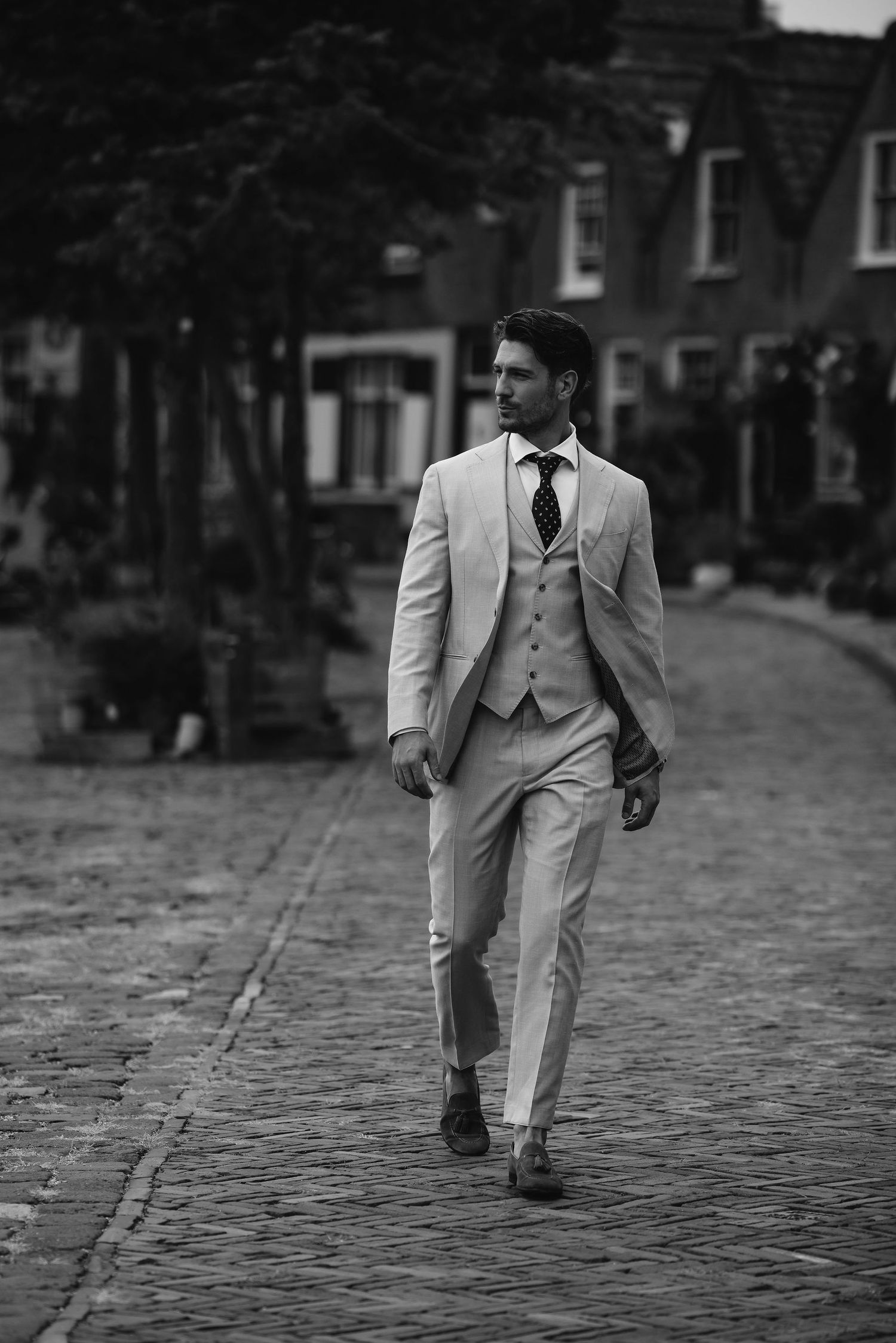 Person wearing light gray three-piece suit walking on cobblestone street in black and white.