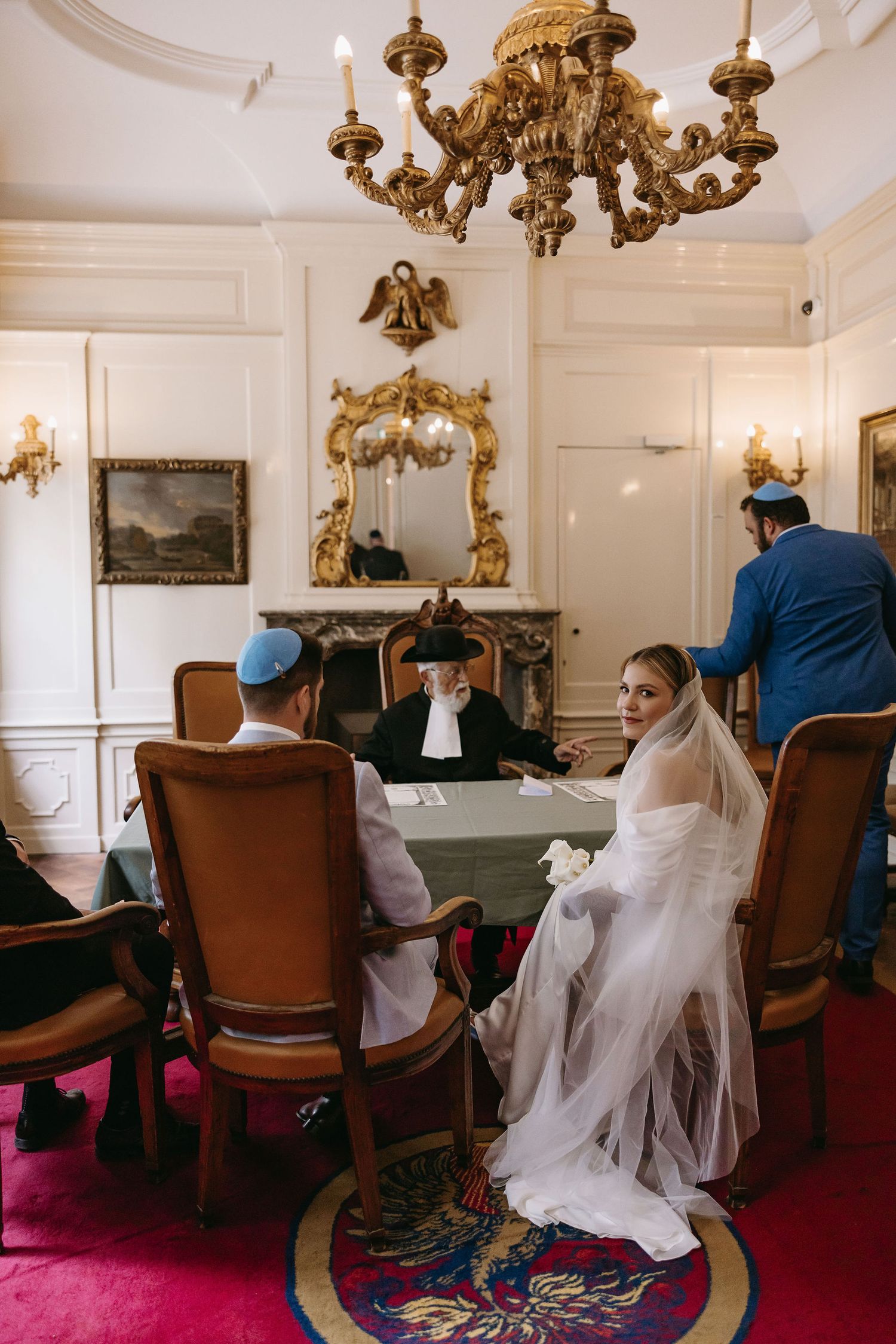 Wedding ceremony in an elegant white room with ornate gold chandeliers and red carpet.