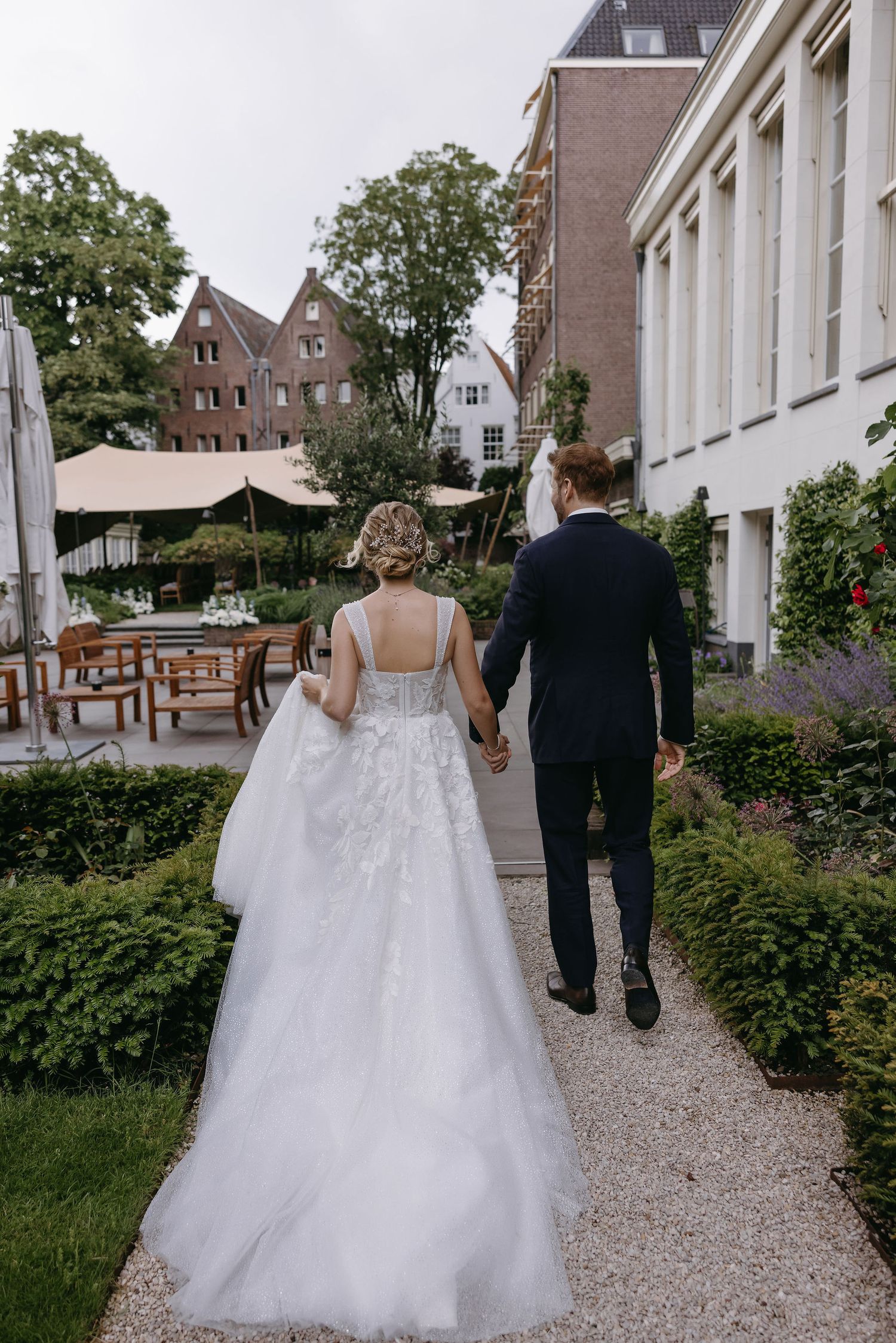 A couple walks hand in hand along a gravel path surrounded by European-style architecture.