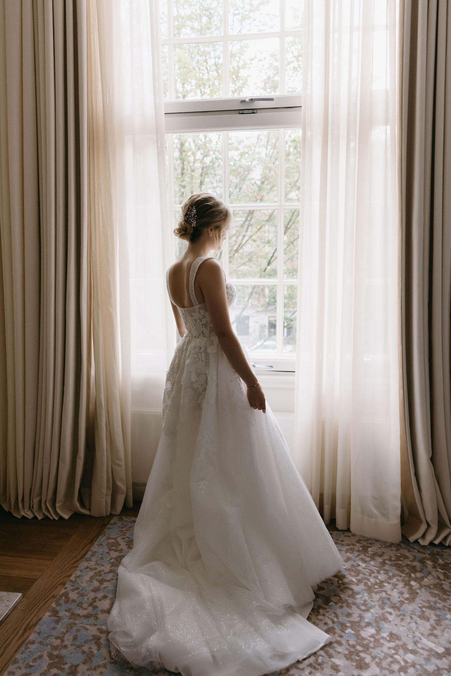 A white wedding dress silhouetted against a bright window with flowing curtains in an elegant room.