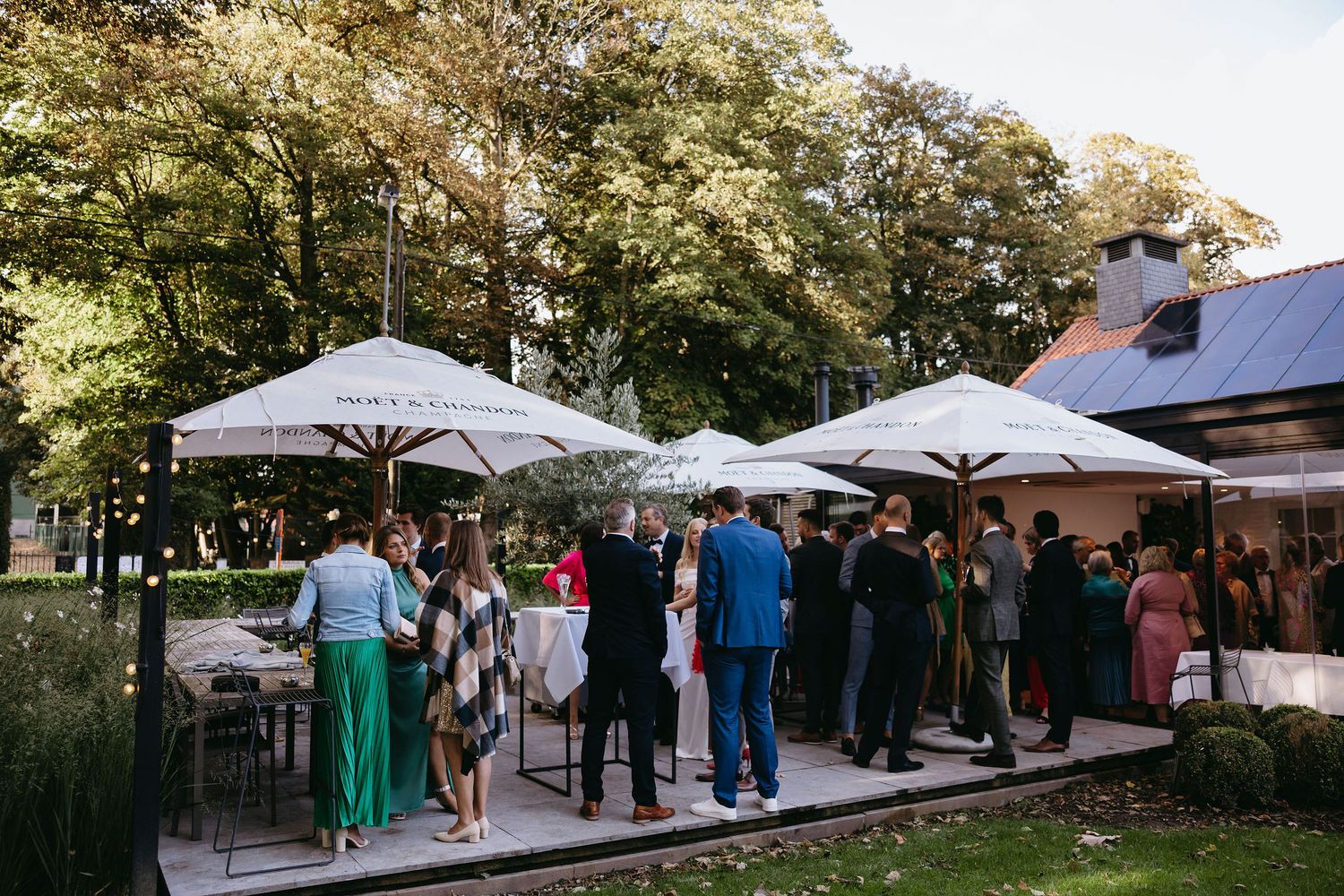 Outdoor wedding reception setup with white umbrellas and guests mingling in a garden setting.