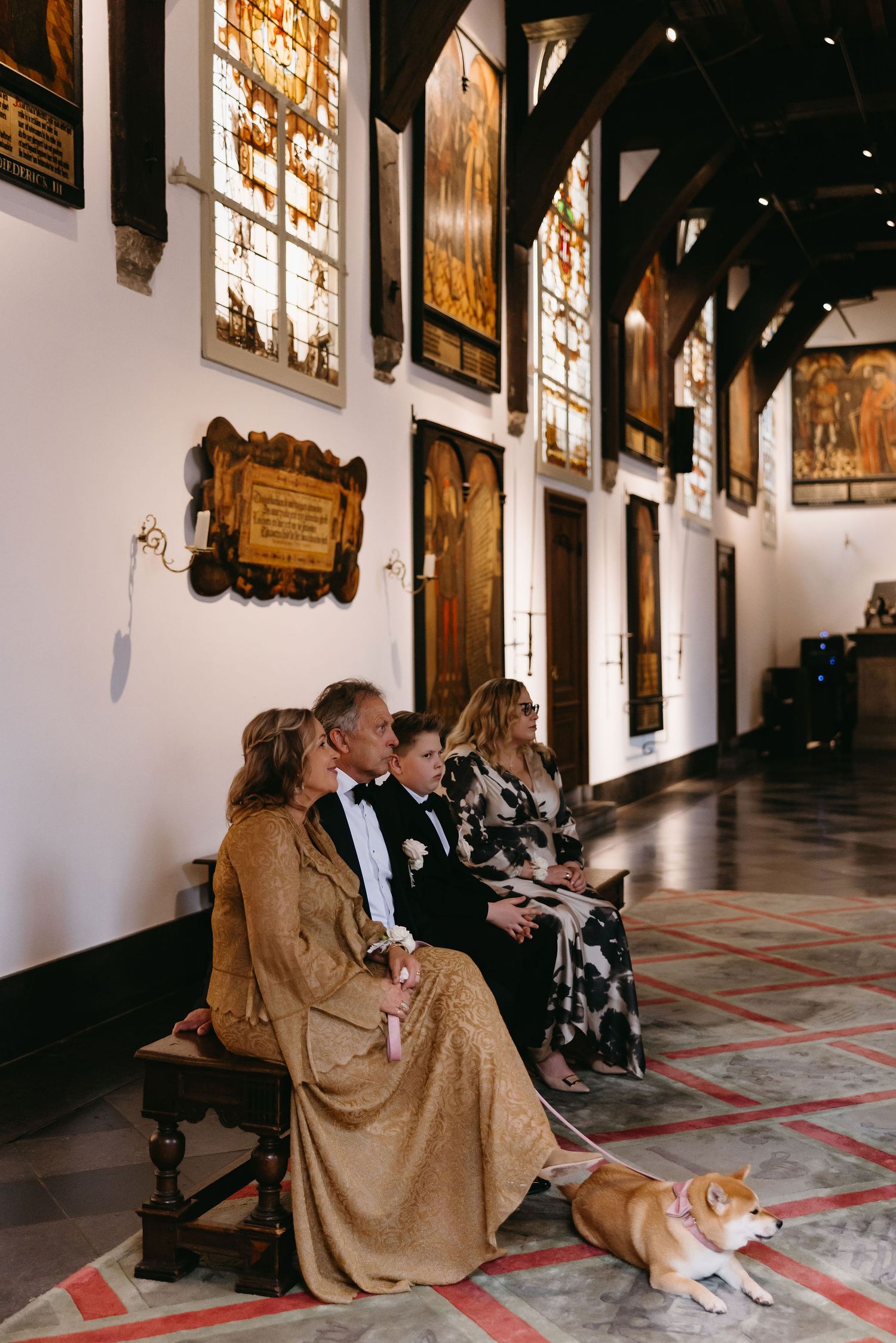 Wedding guests seated in ornate church hall with stained glass windows and a corgi dog resting on floor.