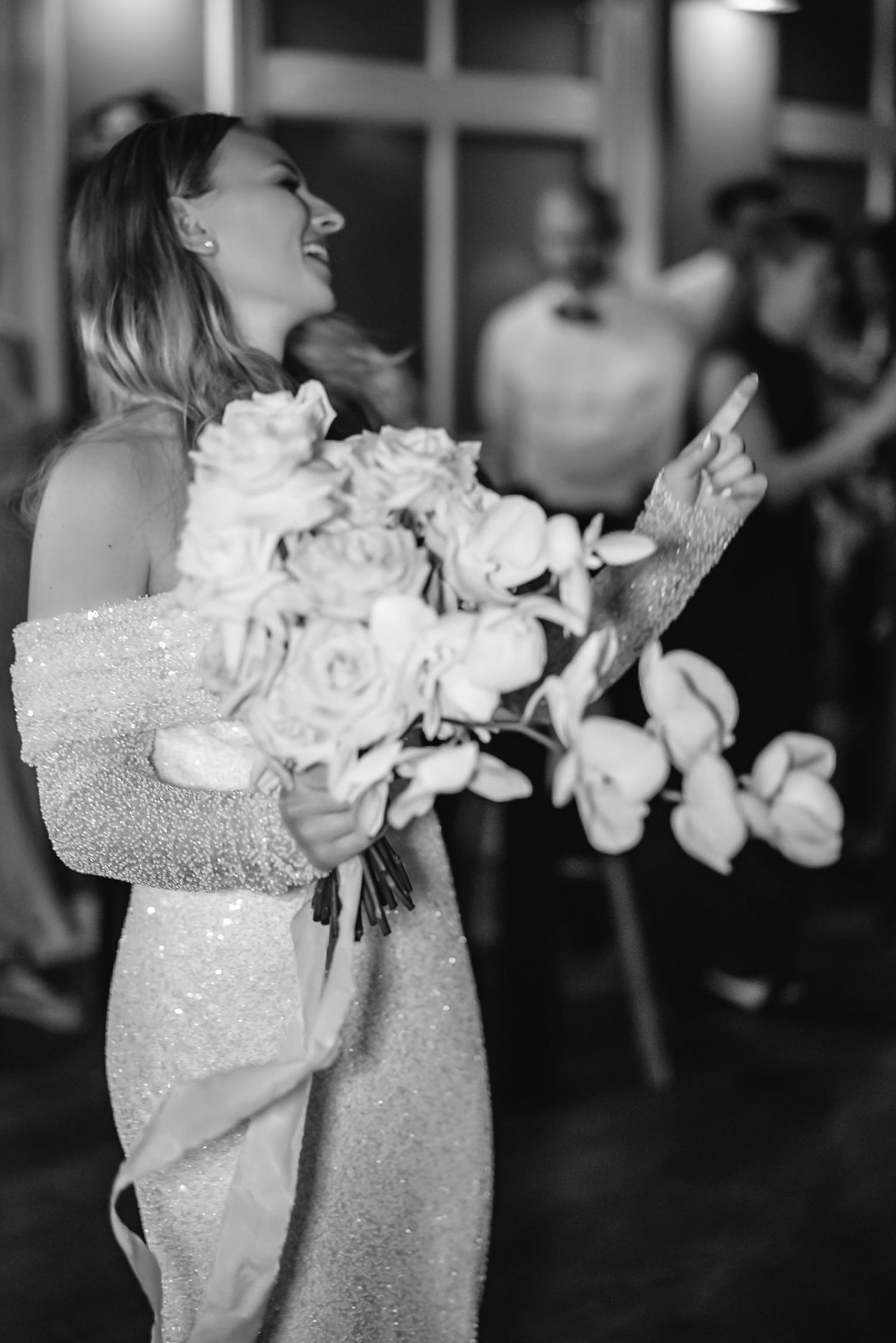 A joyful moment captured in black and white as a bride in a sparkly dress holds a bouquet of white roses and orchids.