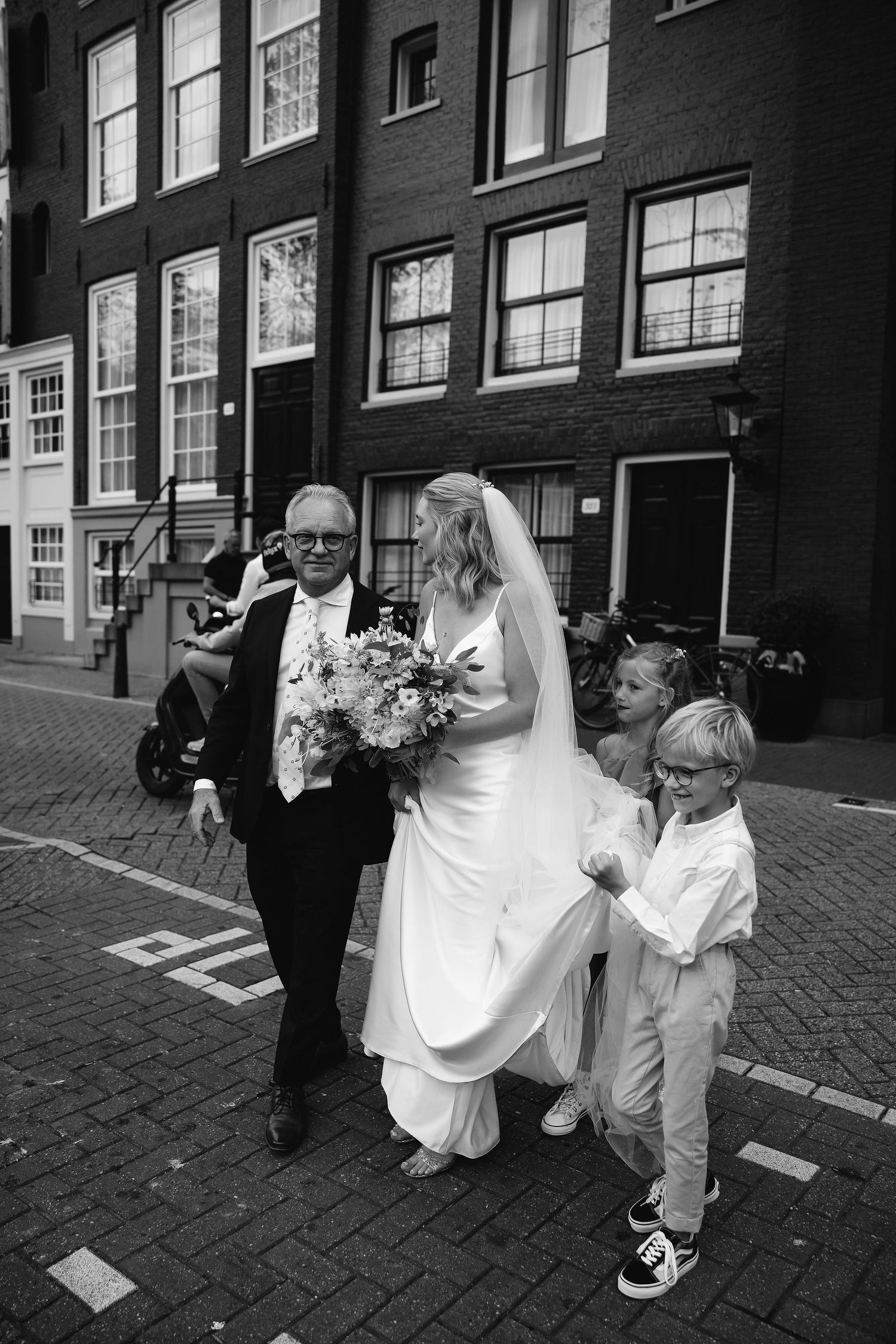 Wedding party walking down Amsterdam street with brick buildings and cobblestones.