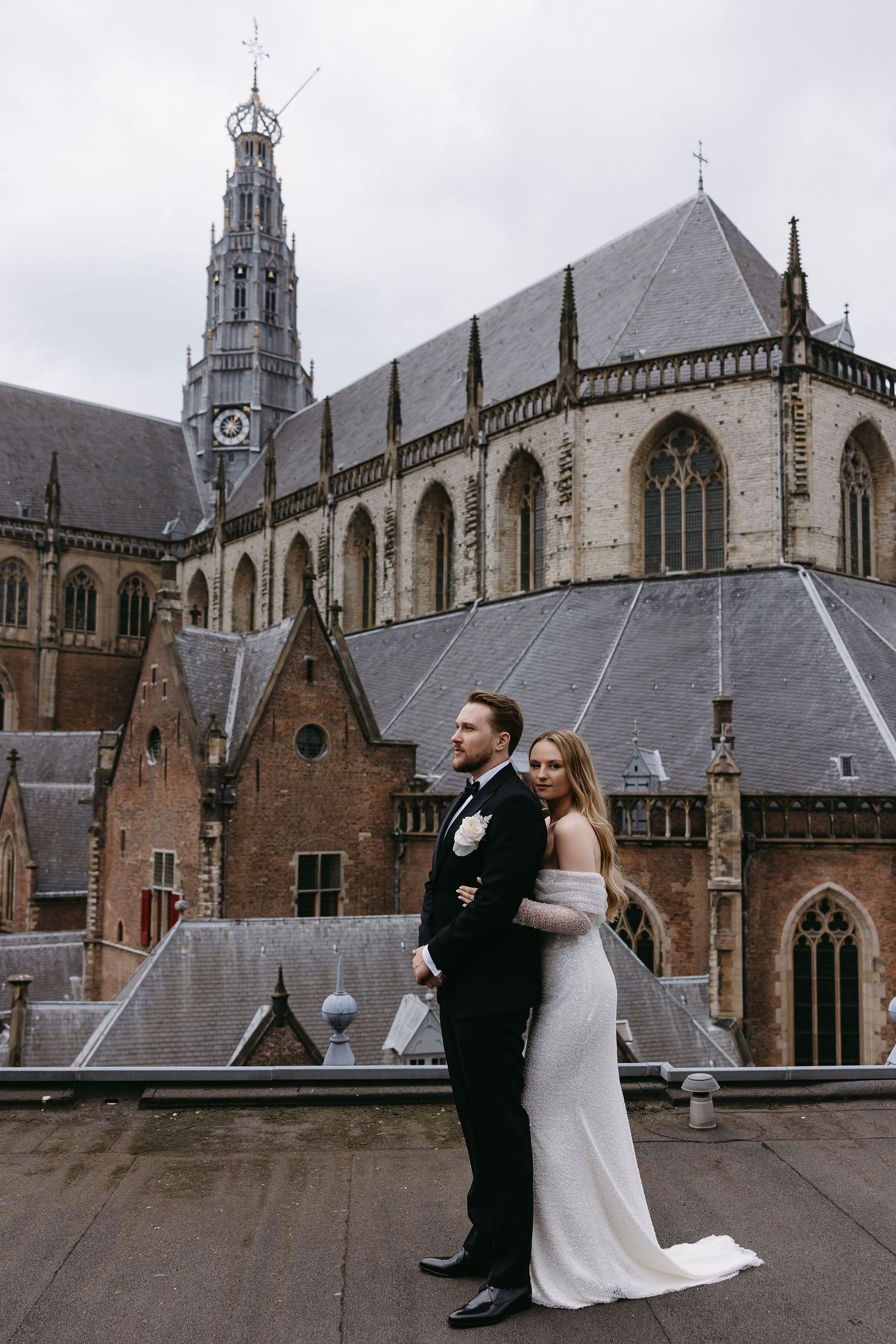 A couple stands on the rooftop of a historic Gothic church with towering spires and arched windows in the background.