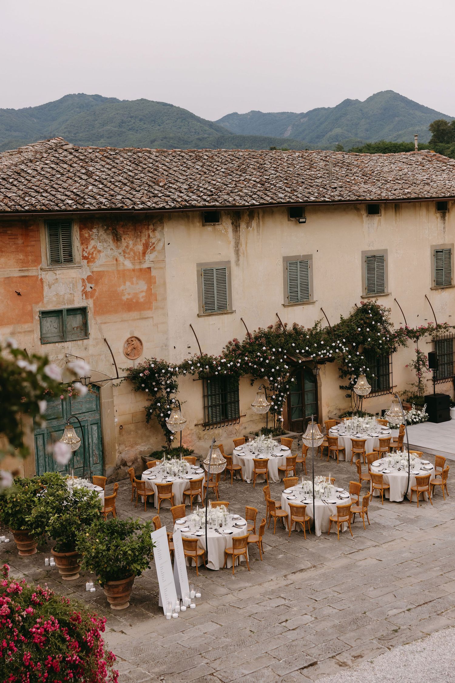 An outdoor Italian wedding reception setup with rustic tables and string lights against a weathered stone building.