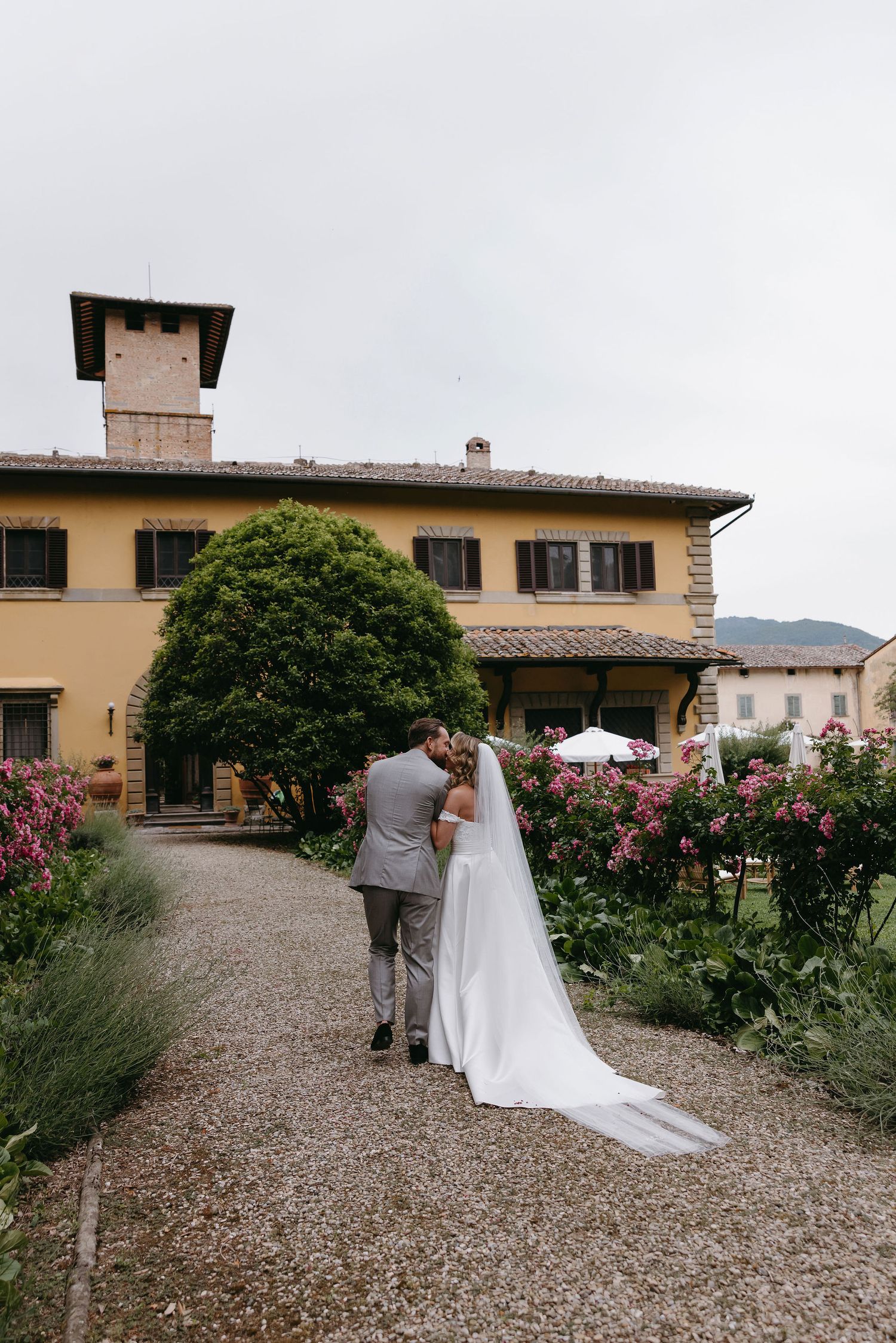 A couple walks along a gravel path beside rose gardens at an Italian villa with beige walls and brown shutters.