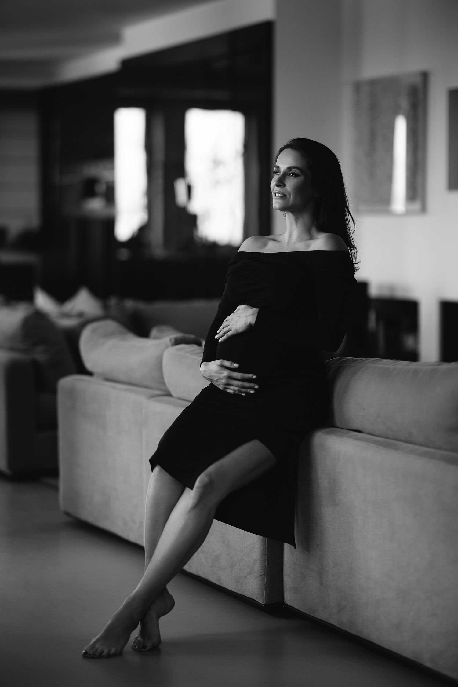 Black and white portrait of a woman in a black dress sitting barefoot on a couch in moody lighting.