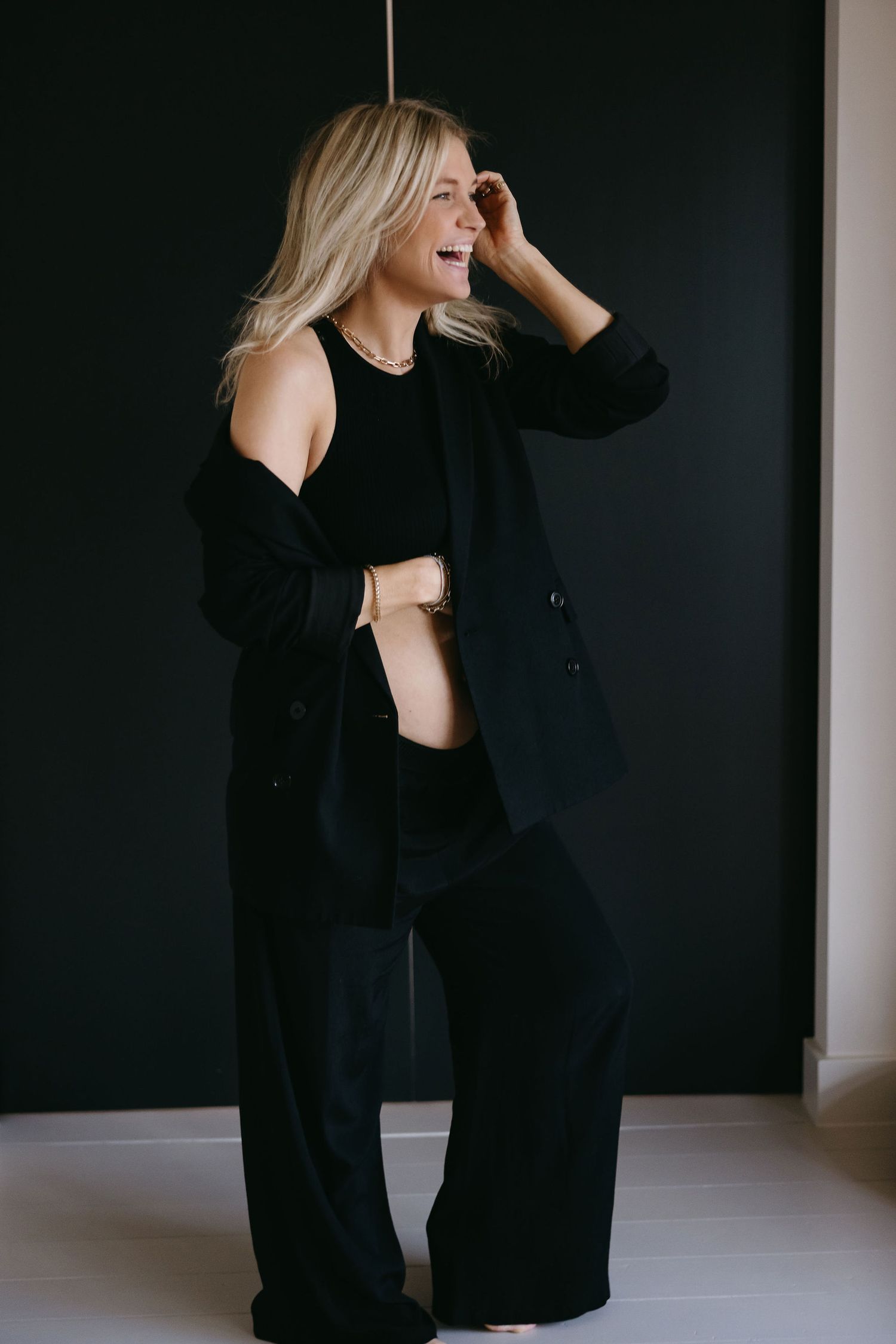 A pregnant woman in a black outfit laughs joyfully against a dark background during a maternity photoshoot.