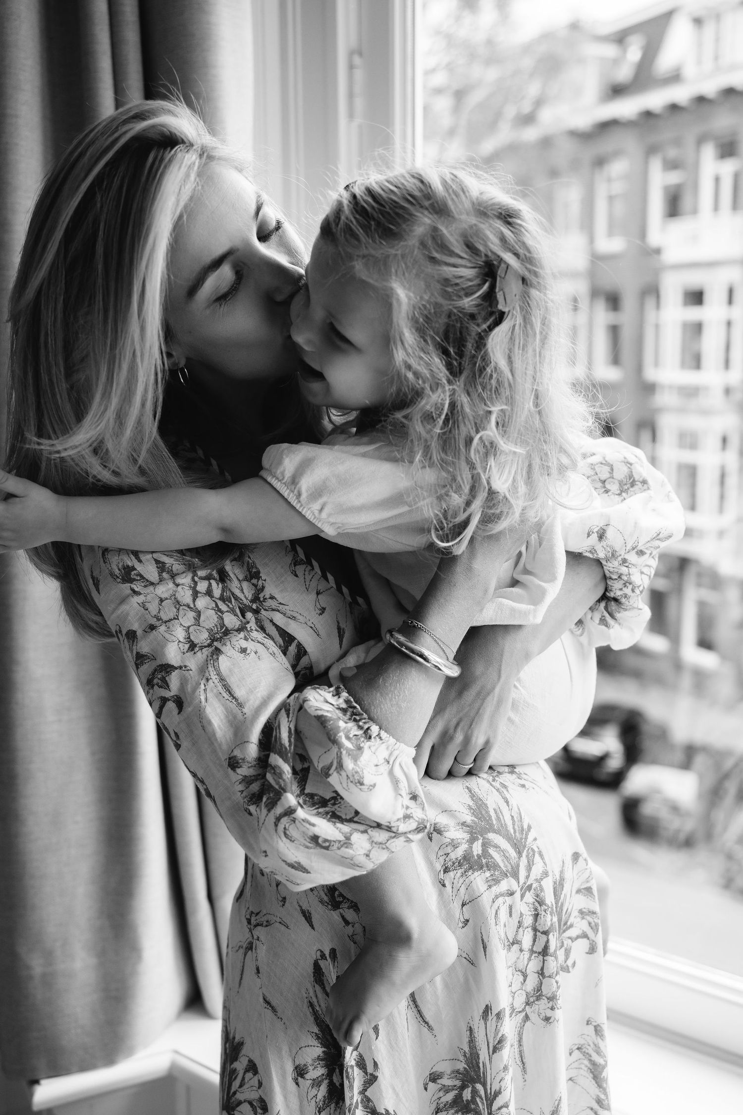 Tender black and white moment between parent and child by window in floral dress.