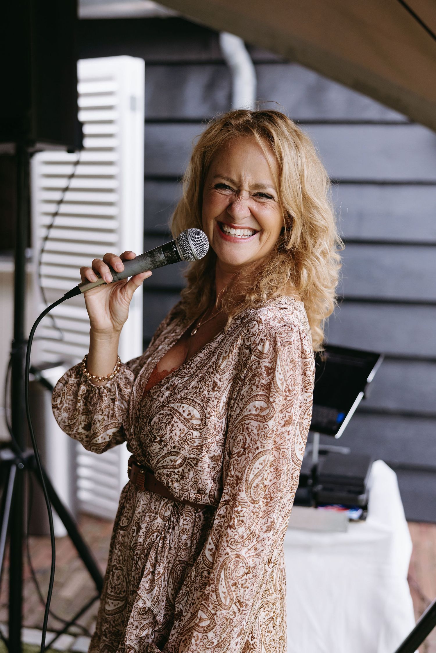 Singer in a patterned paisley dress performs with microphone at outdoor wedding reception.