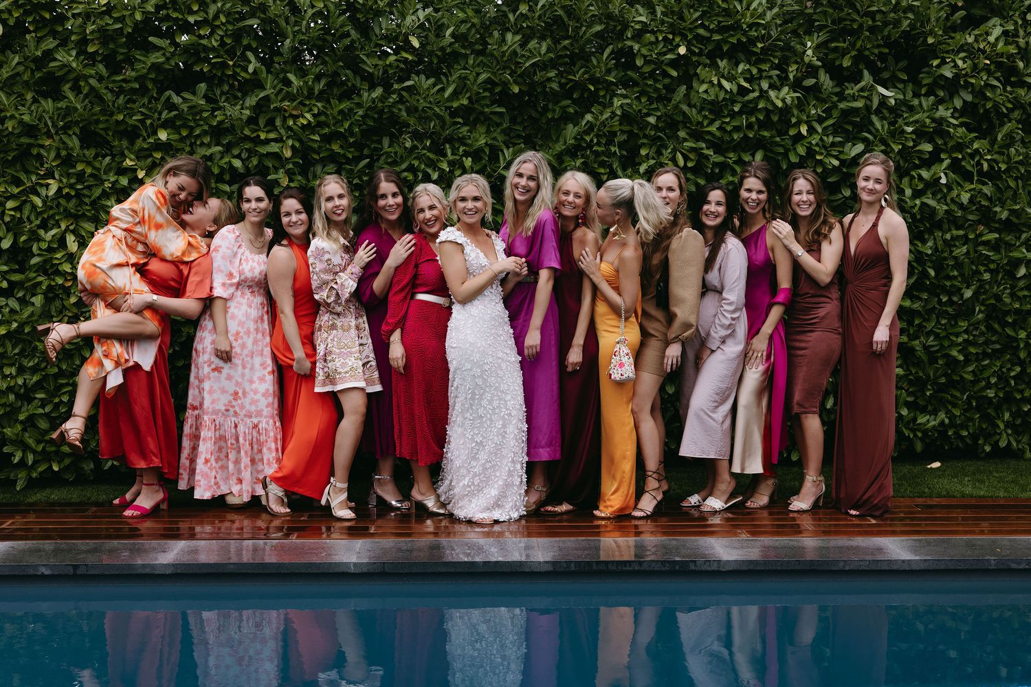 Wedding party in colorful dresses poses by poolside against green hedge backdrop.
