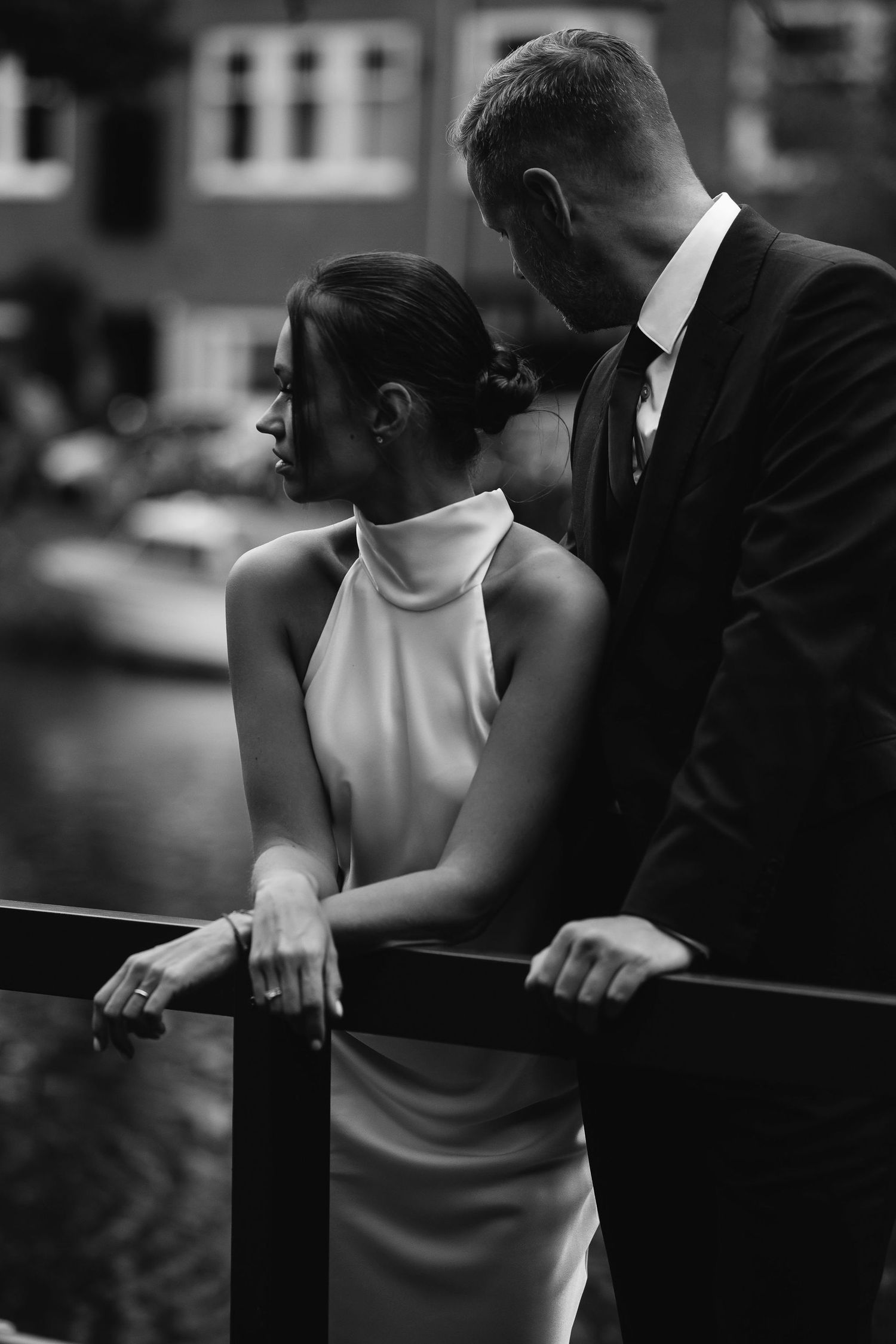 A black and white intimate portrait of a couple in formal attire standing close together against a blurred urban backdrop.