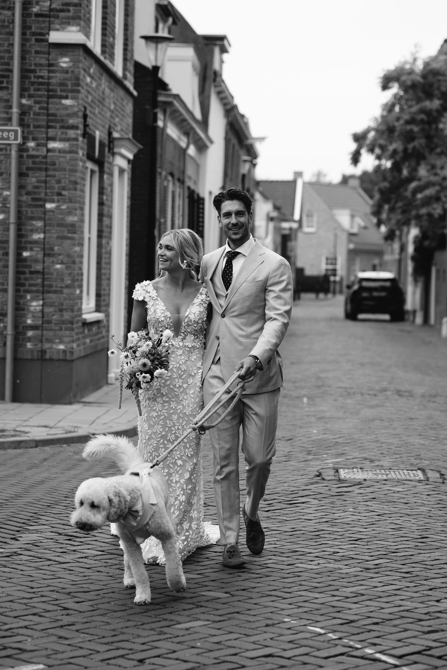 A couple walking their white dog on a cobblestone street lined with brick buildings.