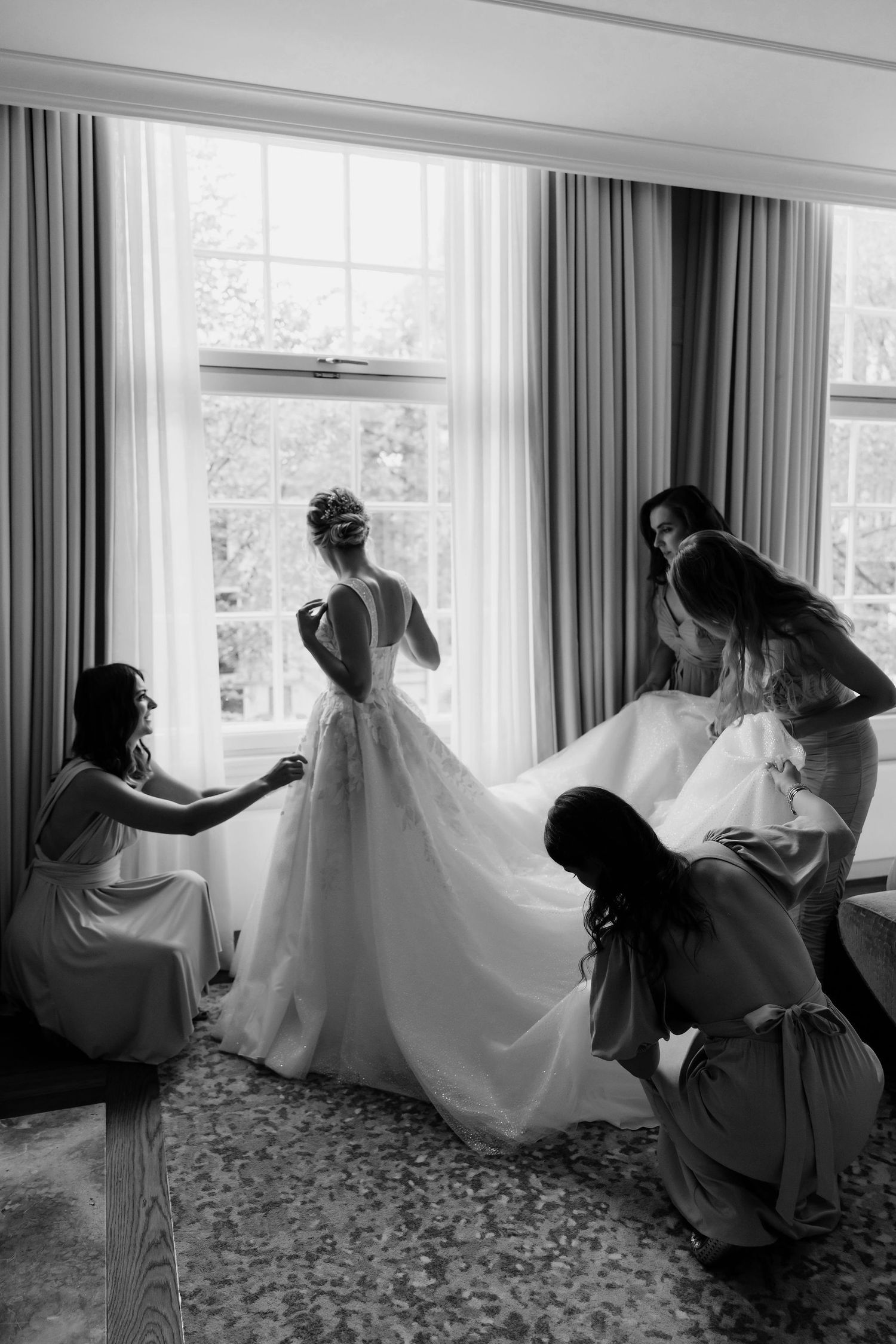 Black and white photograph showing bridesmaids helping adjust a flowing white wedding dress by a bright window.