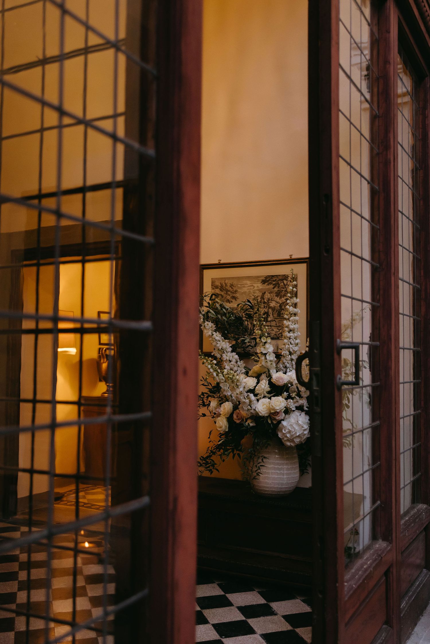 Elegant floral arrangement in a vase seen through a window with checkerboard floor and warm lighting.