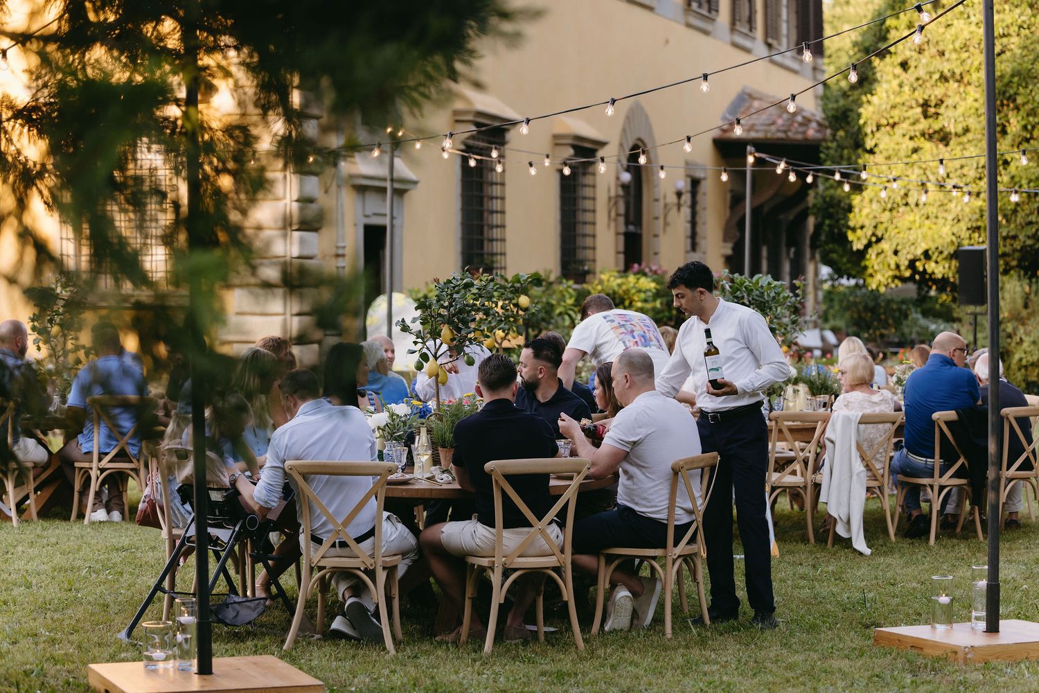 Outdoor wedding reception with guests seated at round tables on a manicured lawn at sunset.