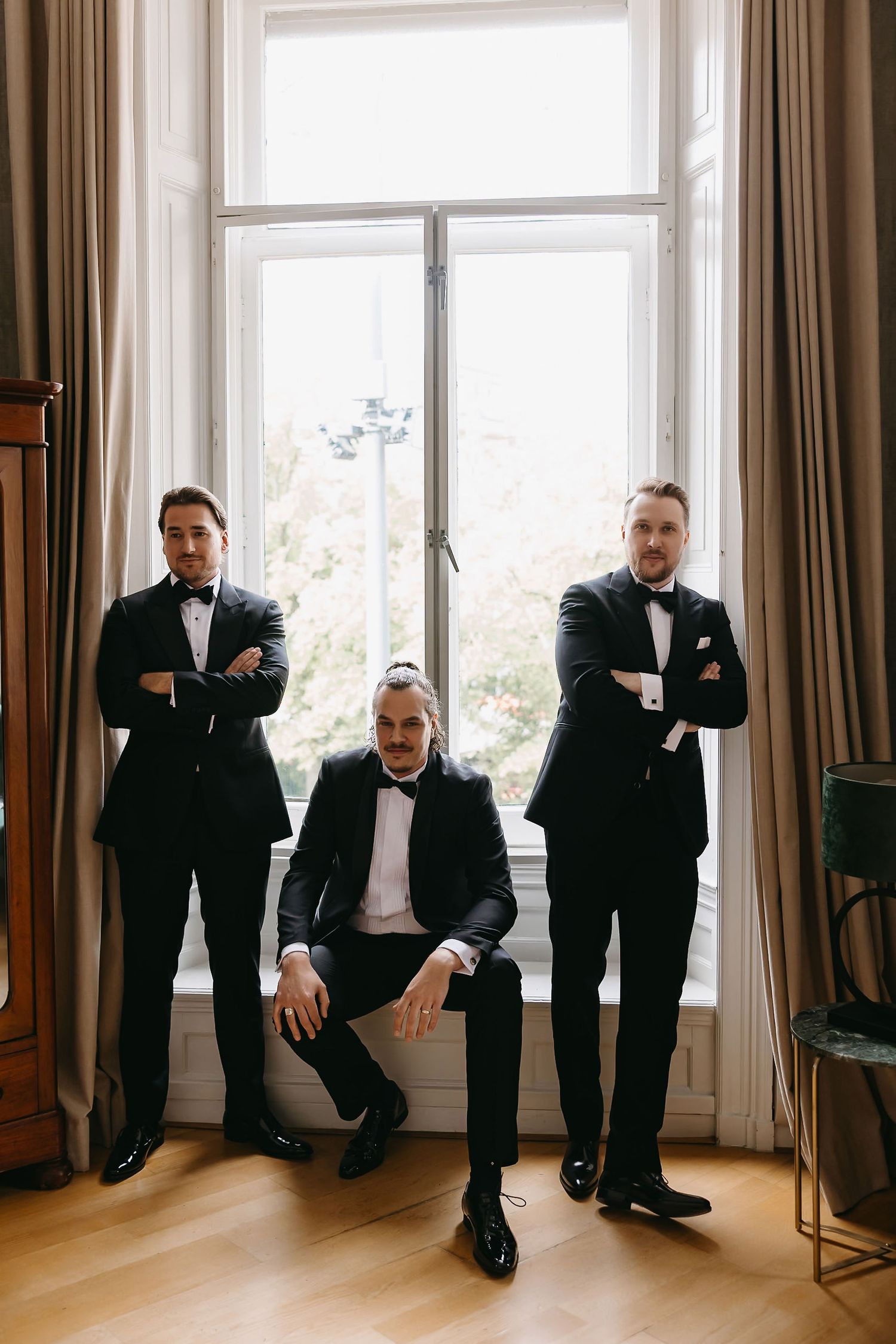 Three groomsmen in black tuxedos pose by bright window in elegant room.