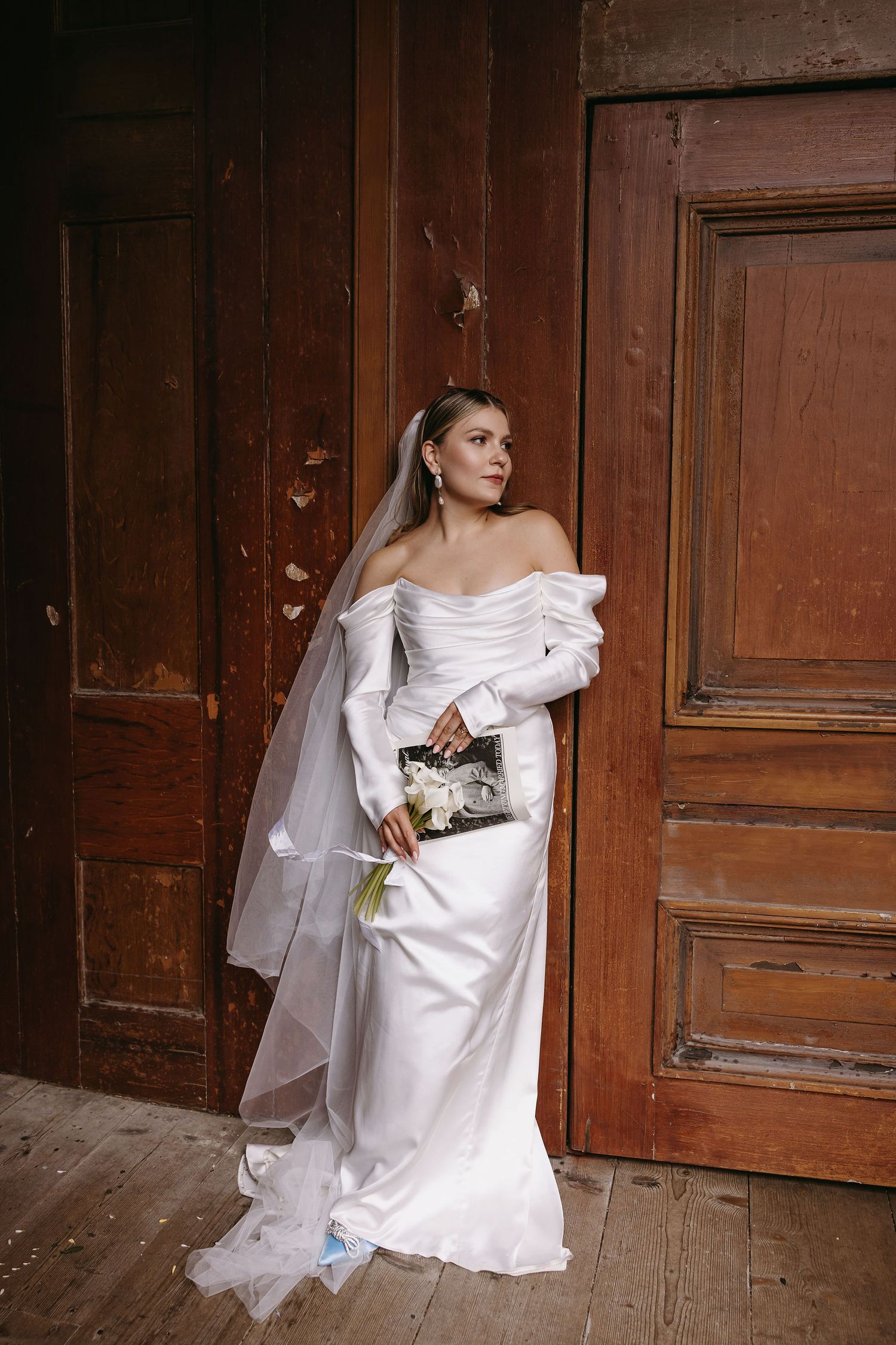 Bride in off-shoulder dress poses against vintage wooden doors with flowing veil.