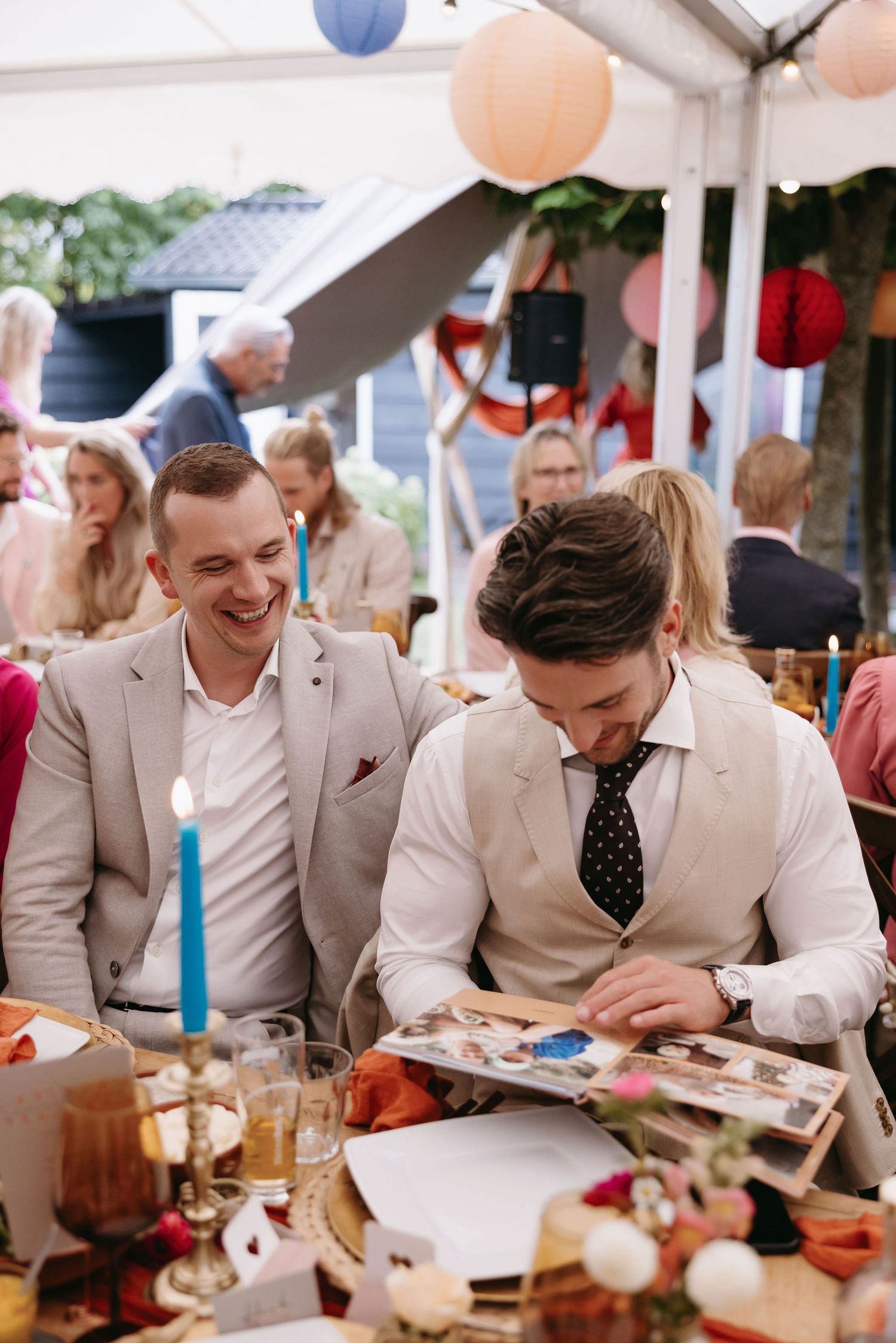 Two guests in light suits seated at decorated wedding reception table with colorful lanterns overhead.