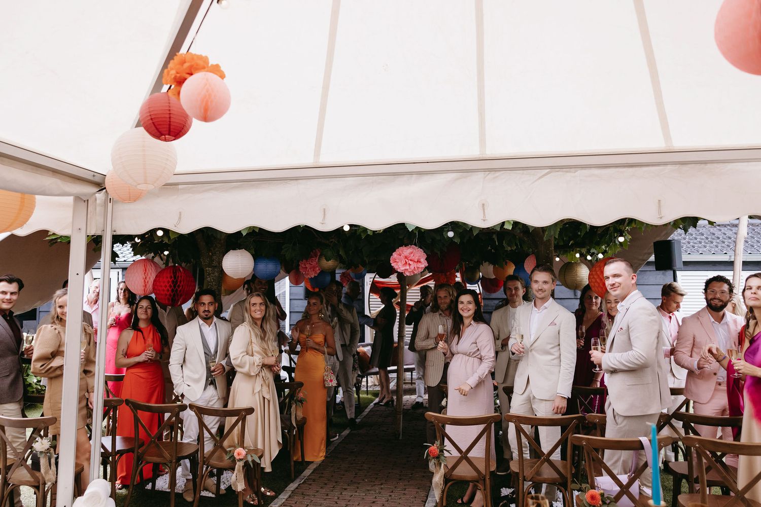 Wedding guests gathered under a white tent decorated with pink lanterns at an outdoor reception.