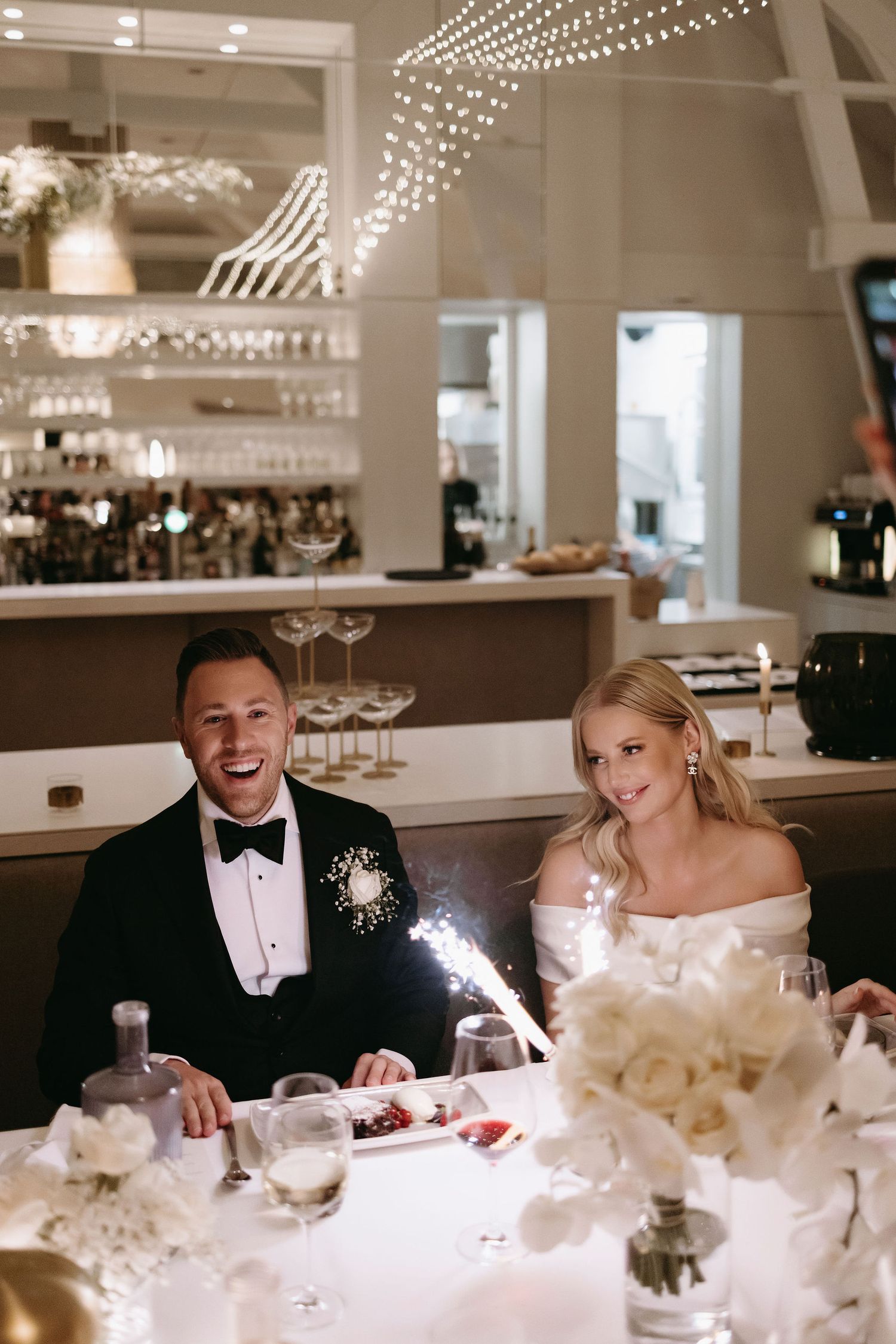Newlyweds share a joyful moment at their reception table decorated with white flowers and glowing sparklers.