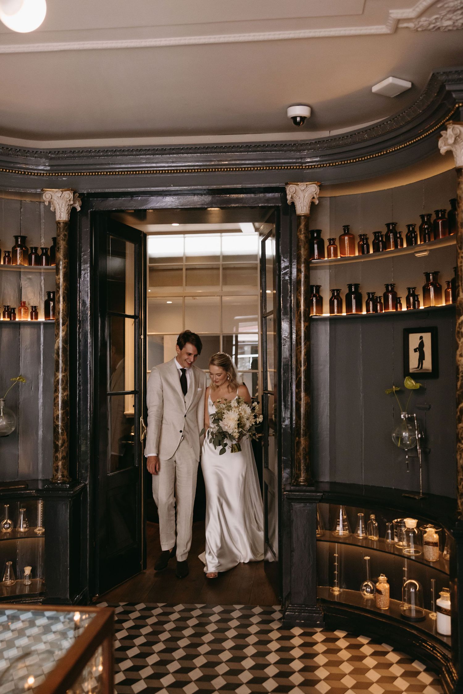 Elegant wedding couple enters through ornate doorway lined with vintage bottles in a sophisticated bar setting.