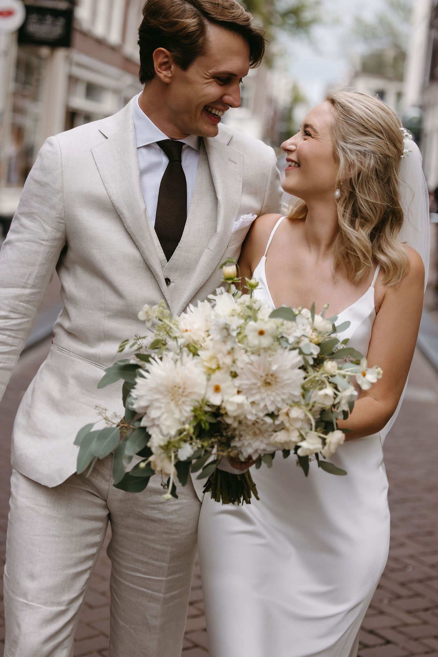 Wedding couple sharing joyful moment on cobblestone street with white floral bouquet.