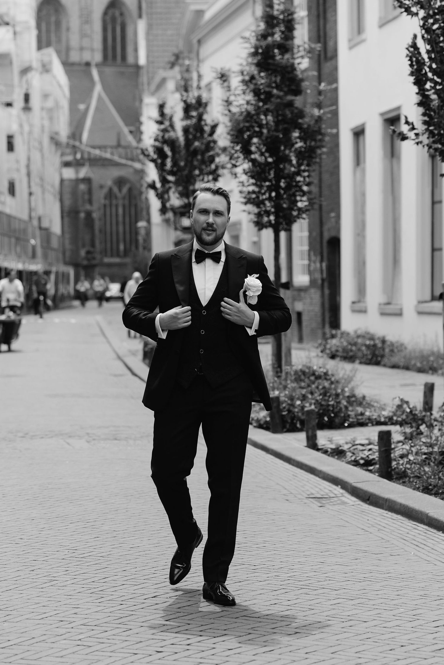 Elegant man in a black tuxedo and bow tie walking down a cobblestone street lined with historic buildings.