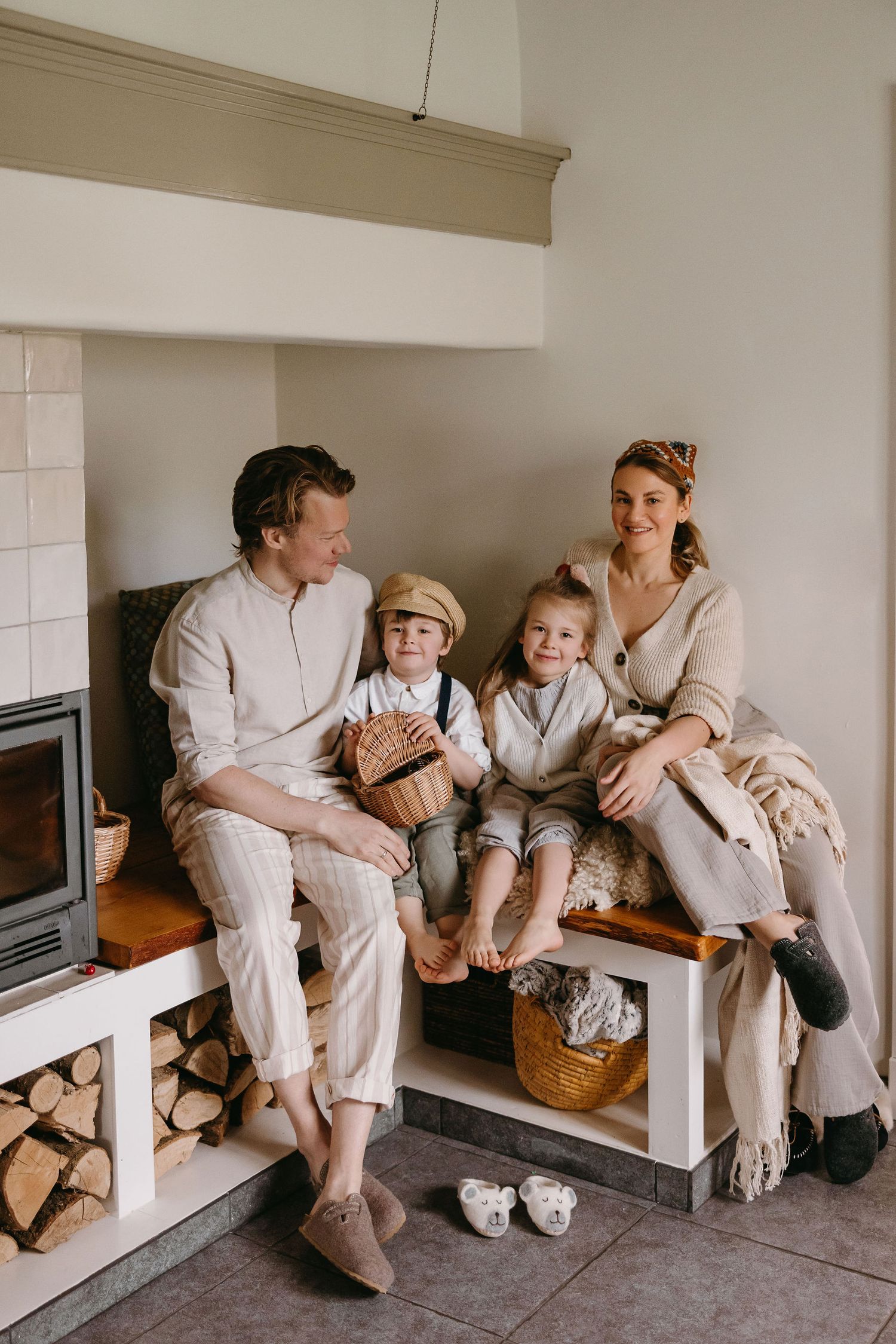 A family in neutral colored clothing sits together on a white bench next to a fireplace with stacked wood.