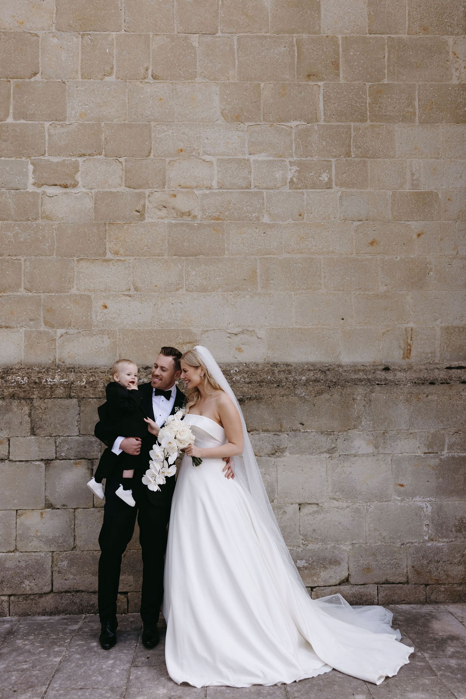 A wedding portrait against a stone wall showcasing an elegant white gown with flowing train.