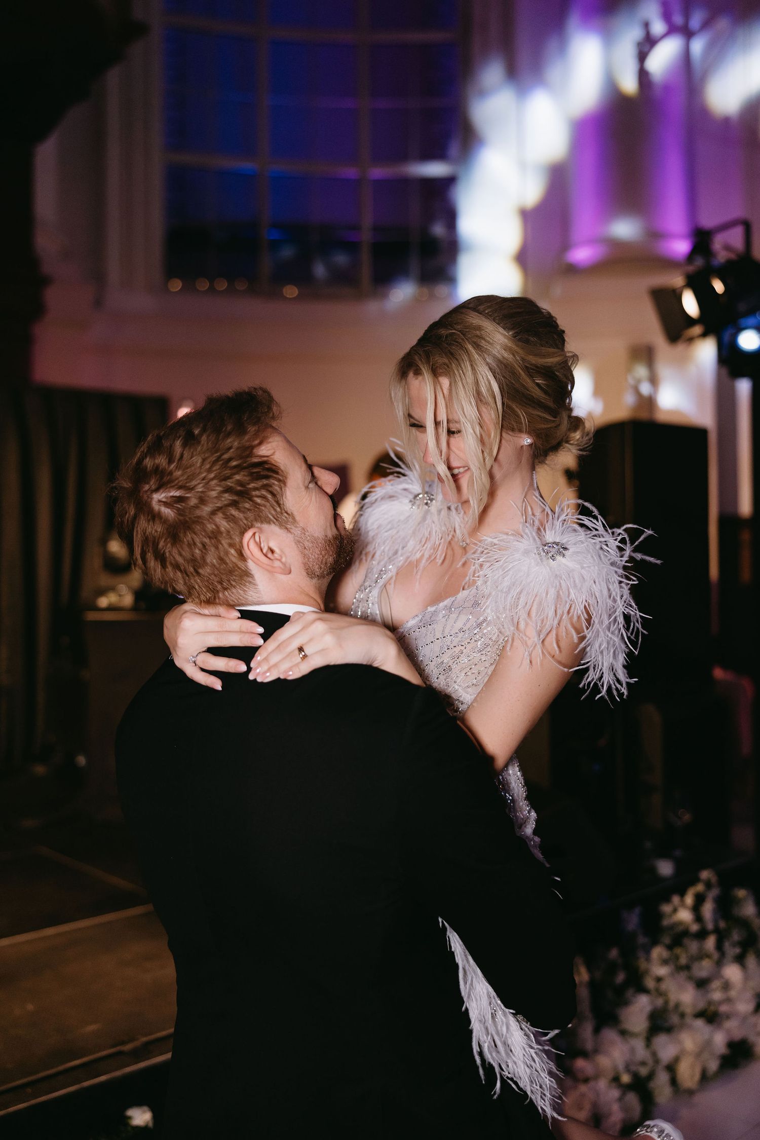 A couple in wedding attire shares a romantic kiss during their evening celebration with purple lighting in the background.