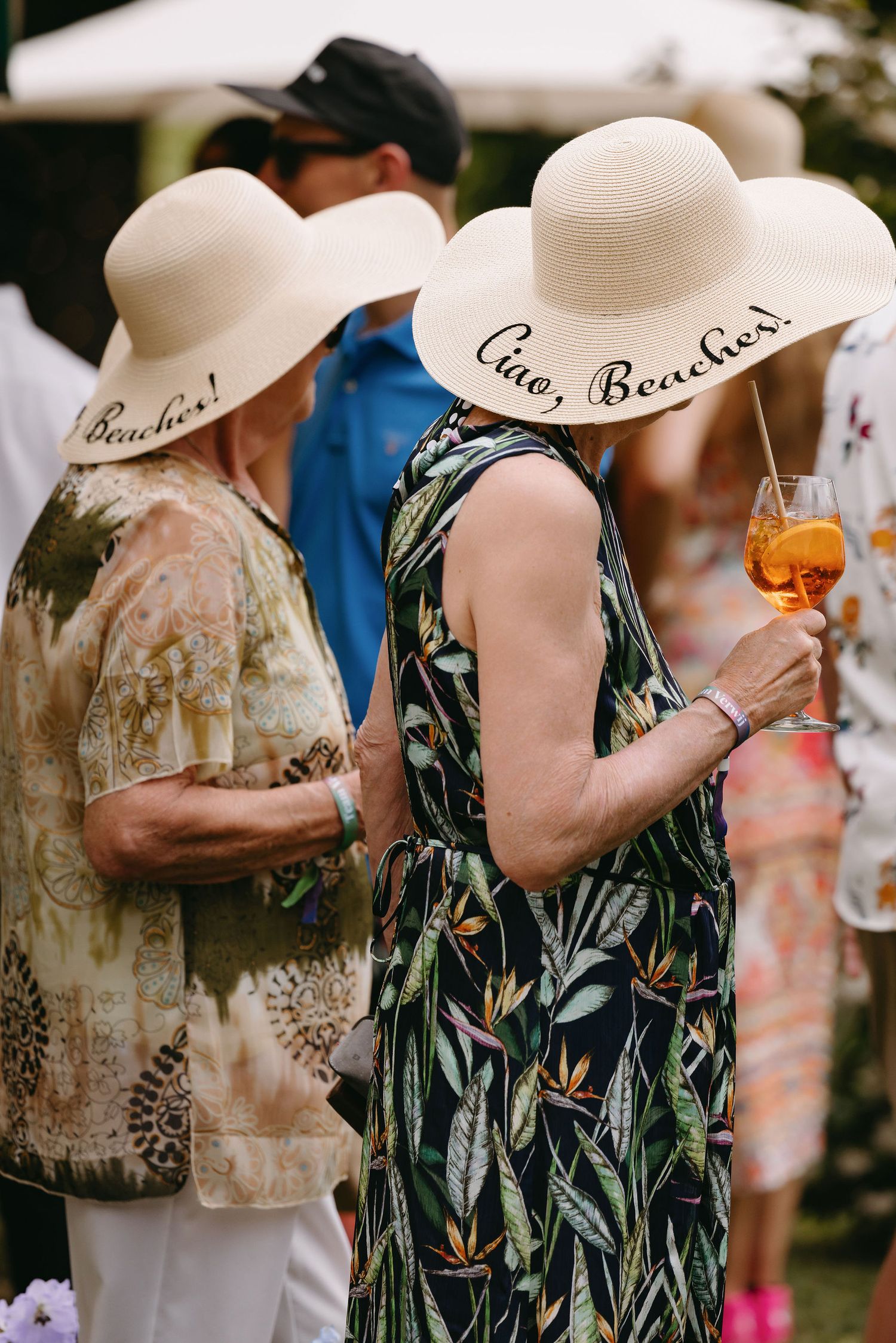 Ladies in summer dresses wearing wide-brimmed straw hats that say 'Ciao Beaches' at an outdoor party.