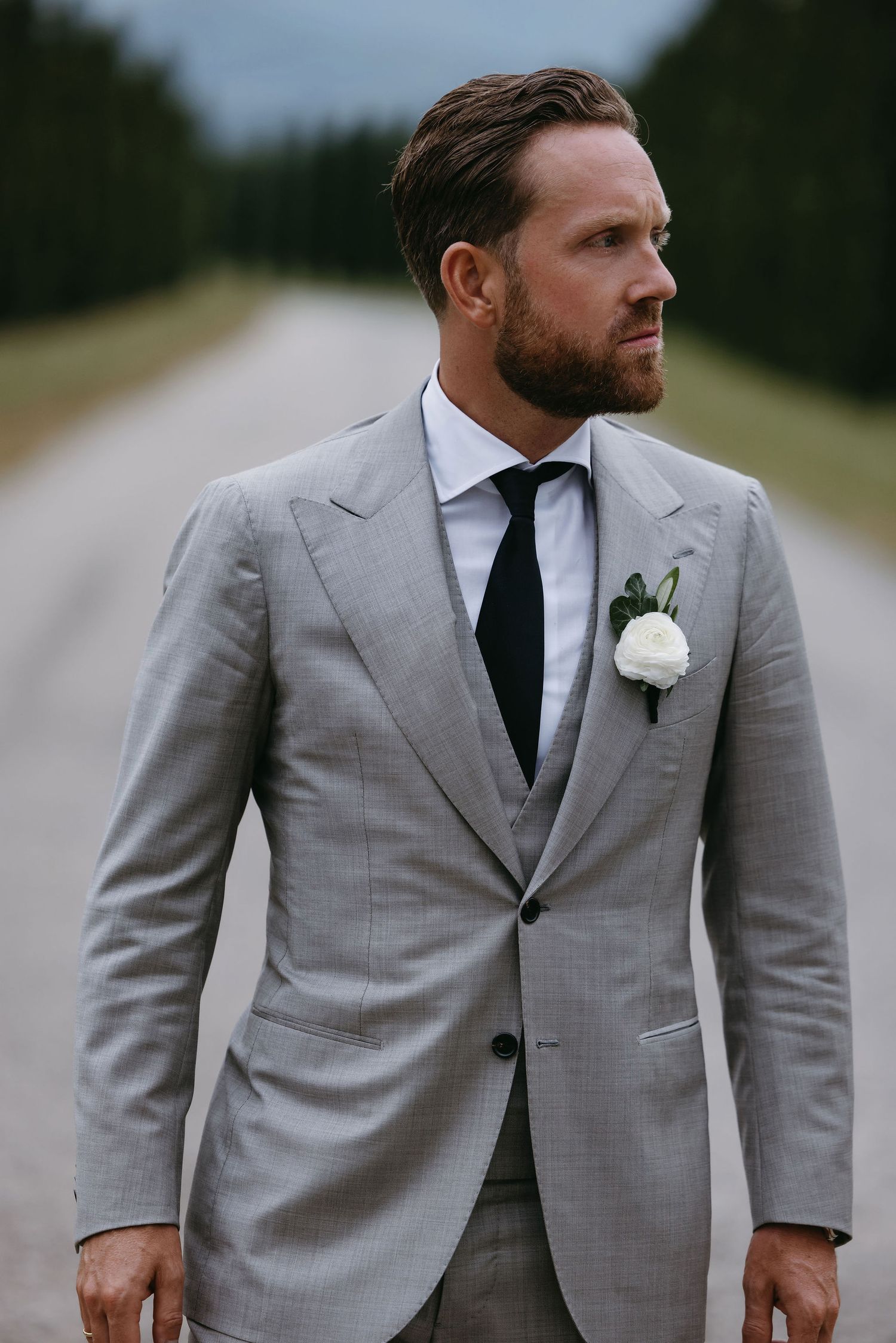 Professional portrait of person wearing light grey suit with white boutonniere against rural background.