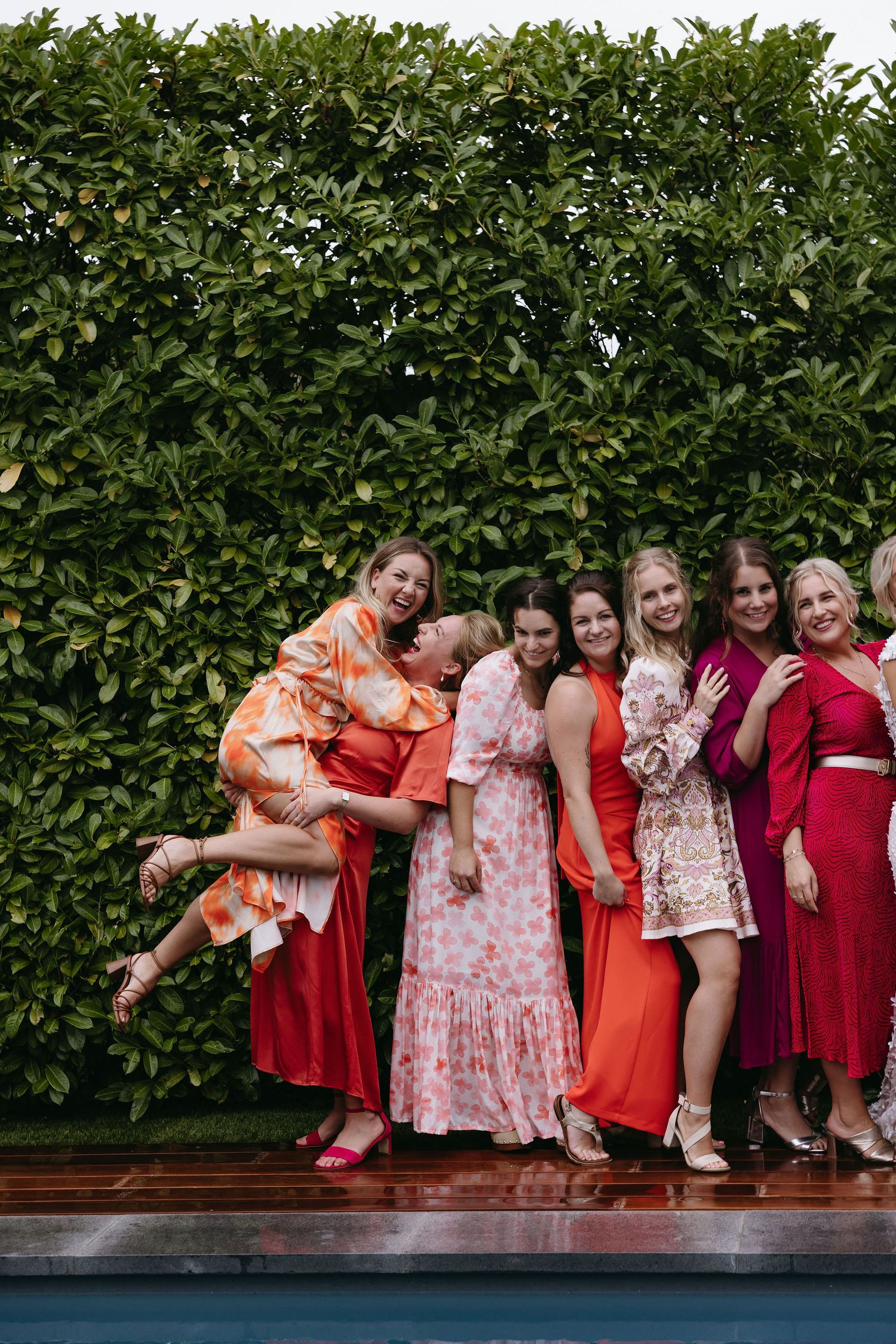 Bridal party in shades of red and pink poses playfully against a green hedge.